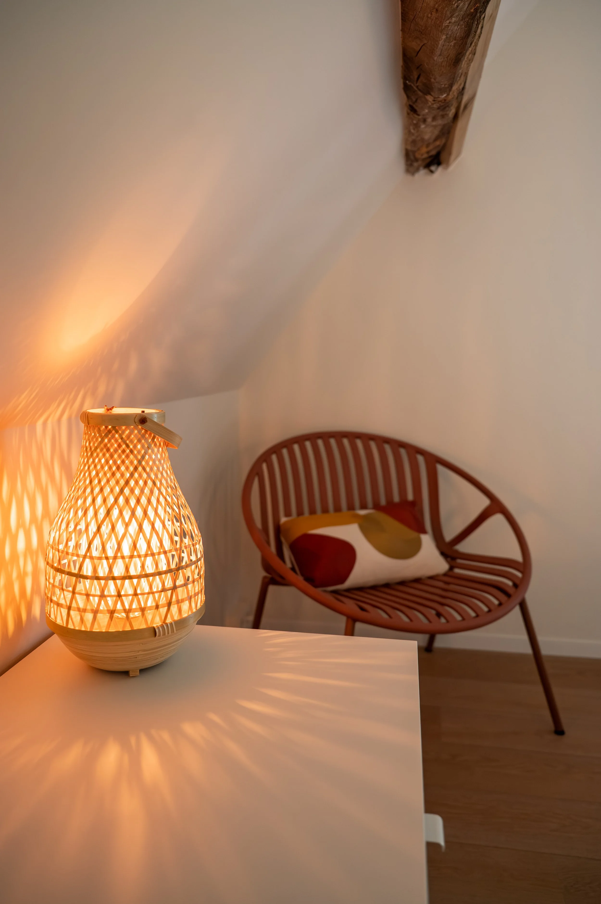 A warmly lit table lamp with a lattice design casts shadows on a nearby white surface. In the background, there is a wooden bench with a colorful cushion and a wall with a visible wooden beam.