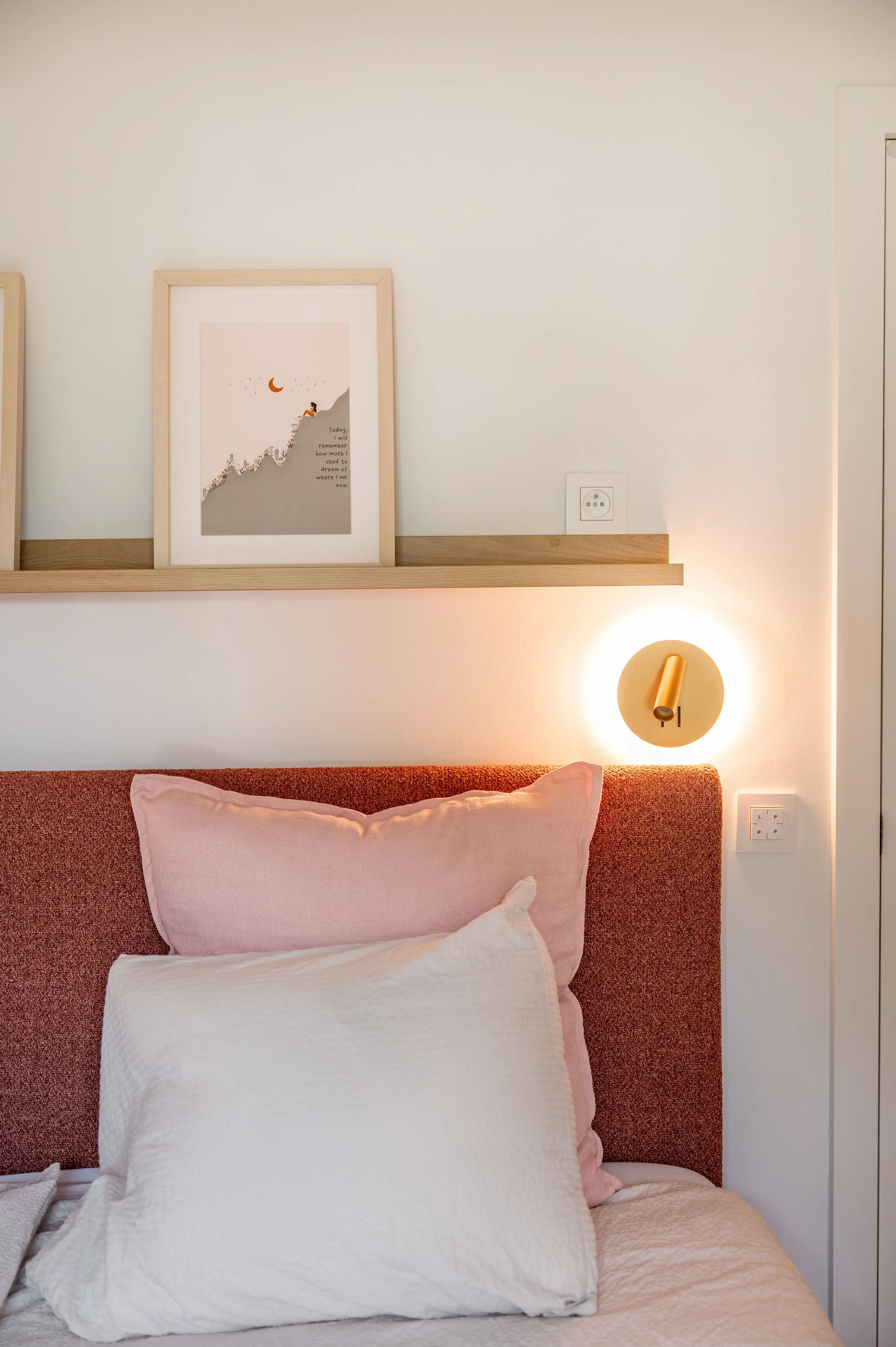 A bedroom with a bed that has a pink pillow and white bedspread. Behind the bed, there is a light-colored wall with framed artwork and a wall-mounted lamp. The artwork depicts a person sitting on a mountain with a moon and stars, with a quote.