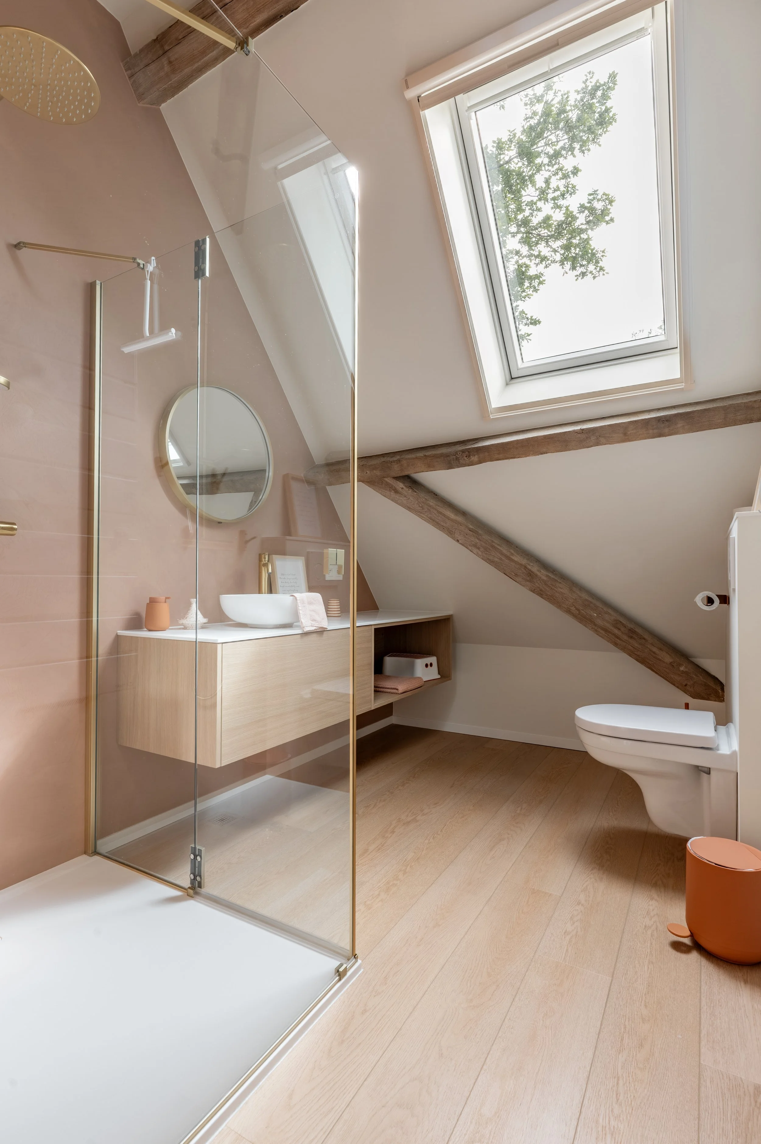 Modern bathroom with a sloped ceiling, wooden beams, skylight window revealing a tree outside, a glass shower enclosure, a floating vanity with a vessel sink, a round mirror, a toilet, and a small orange trash bin.