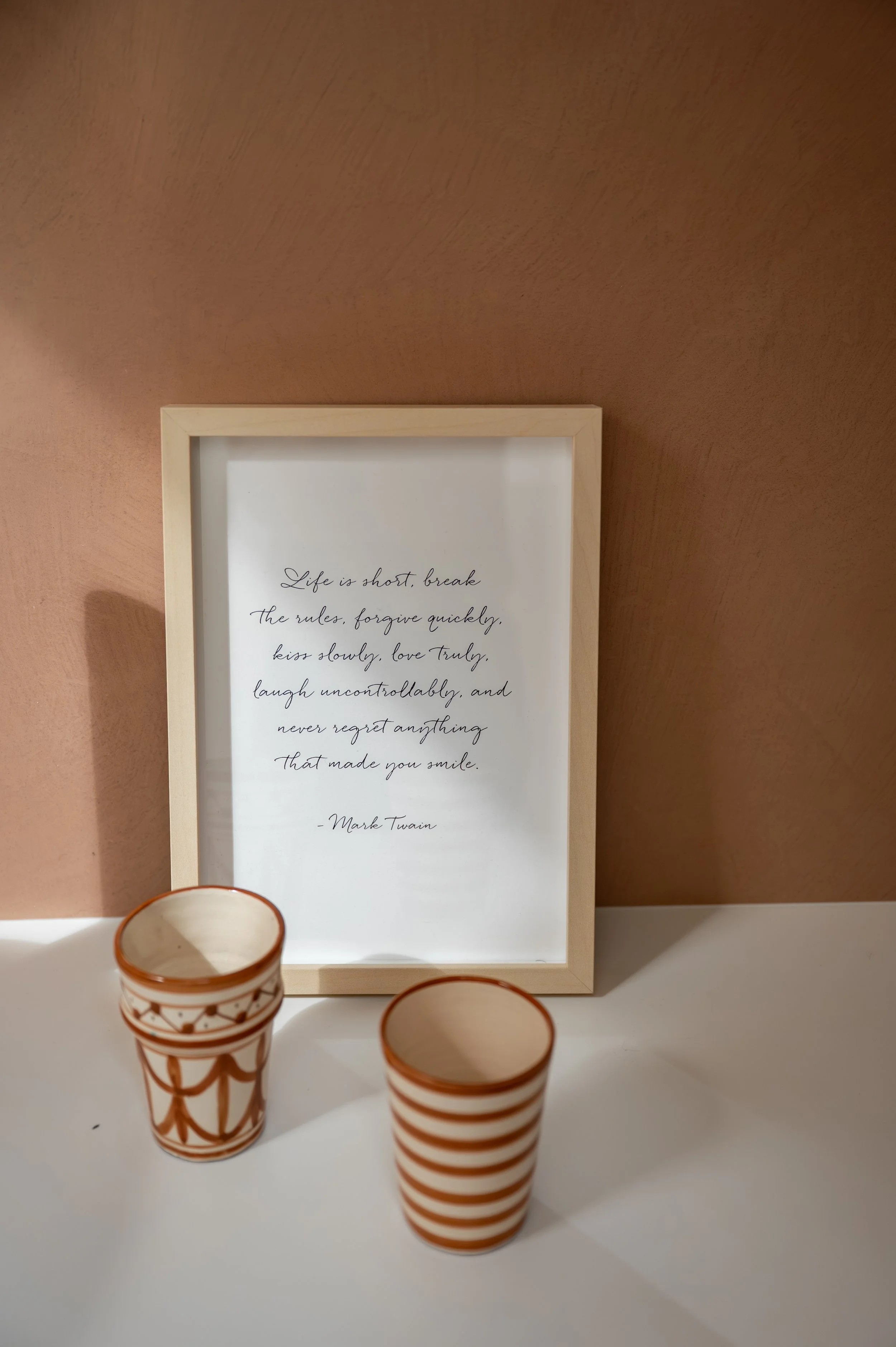 Three striped paper cups on a white surface, a framed quote on a brown wall, and the top of a decorative plant or object in the bottom left corner.