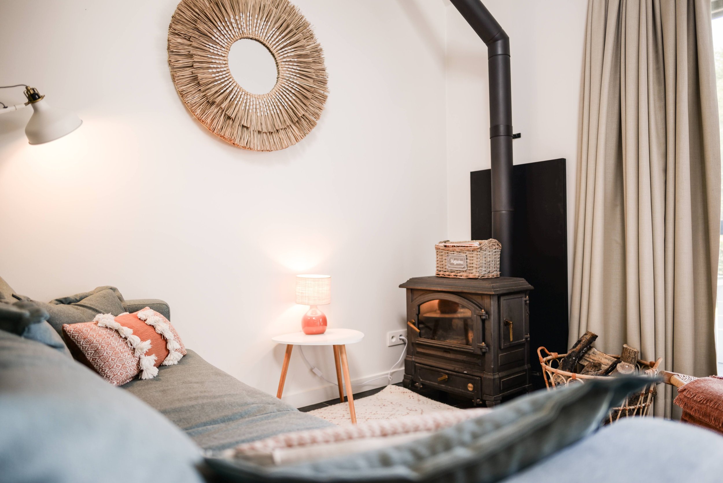 A cozy living room corner featuring a gray sofa with patterned throw pillows, a white side table with a pink lamp, a wicker basket, a black wood stove, and beige curtains. A decorative round mirror made of natural fibers hangs on the wall.