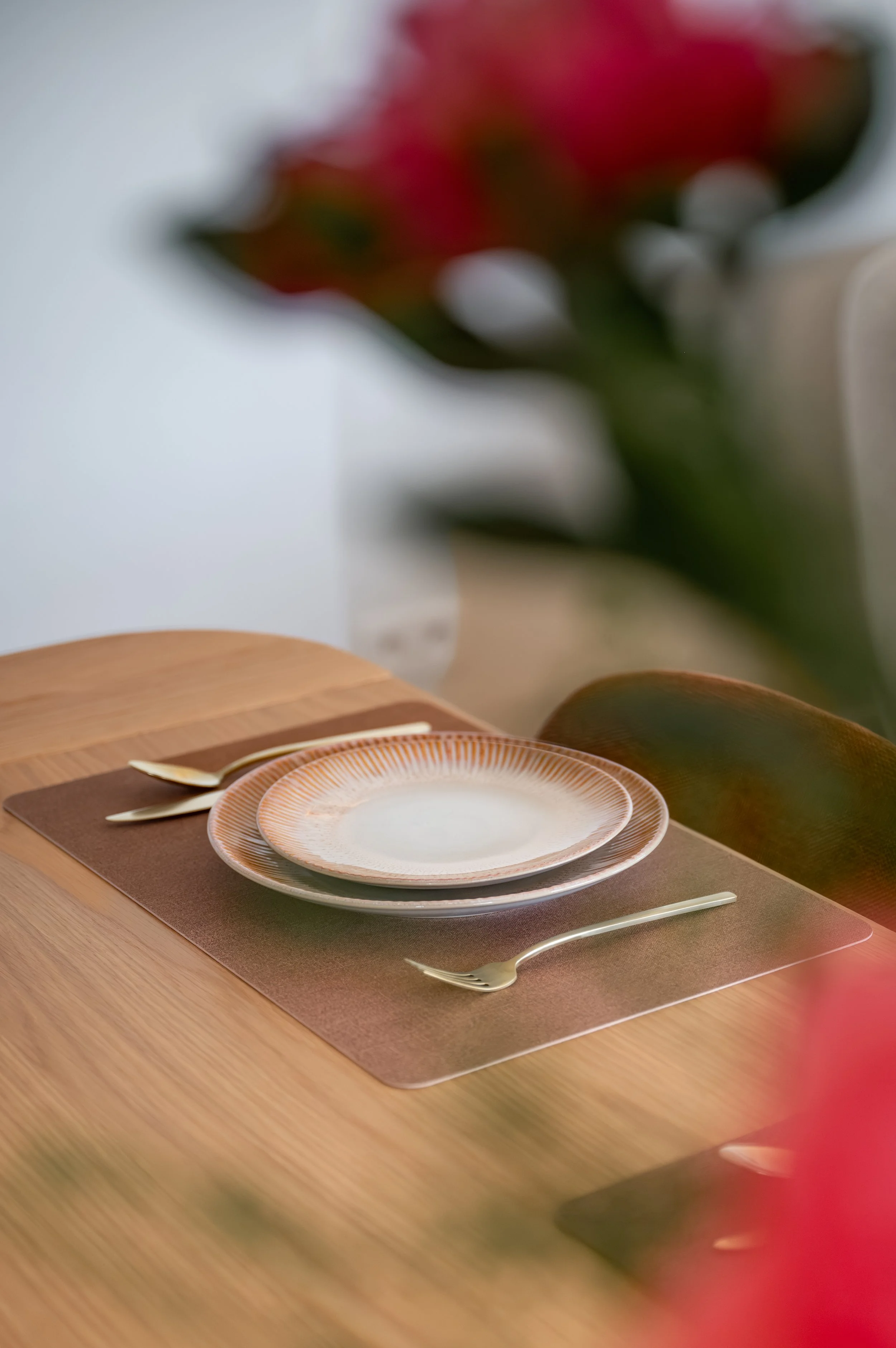 An empty dinner plate with a decorative rim, placed on a placemat on a wooden table, with a matching fork and knife set nearby. In the background, blurred floral arrangement and a wall.