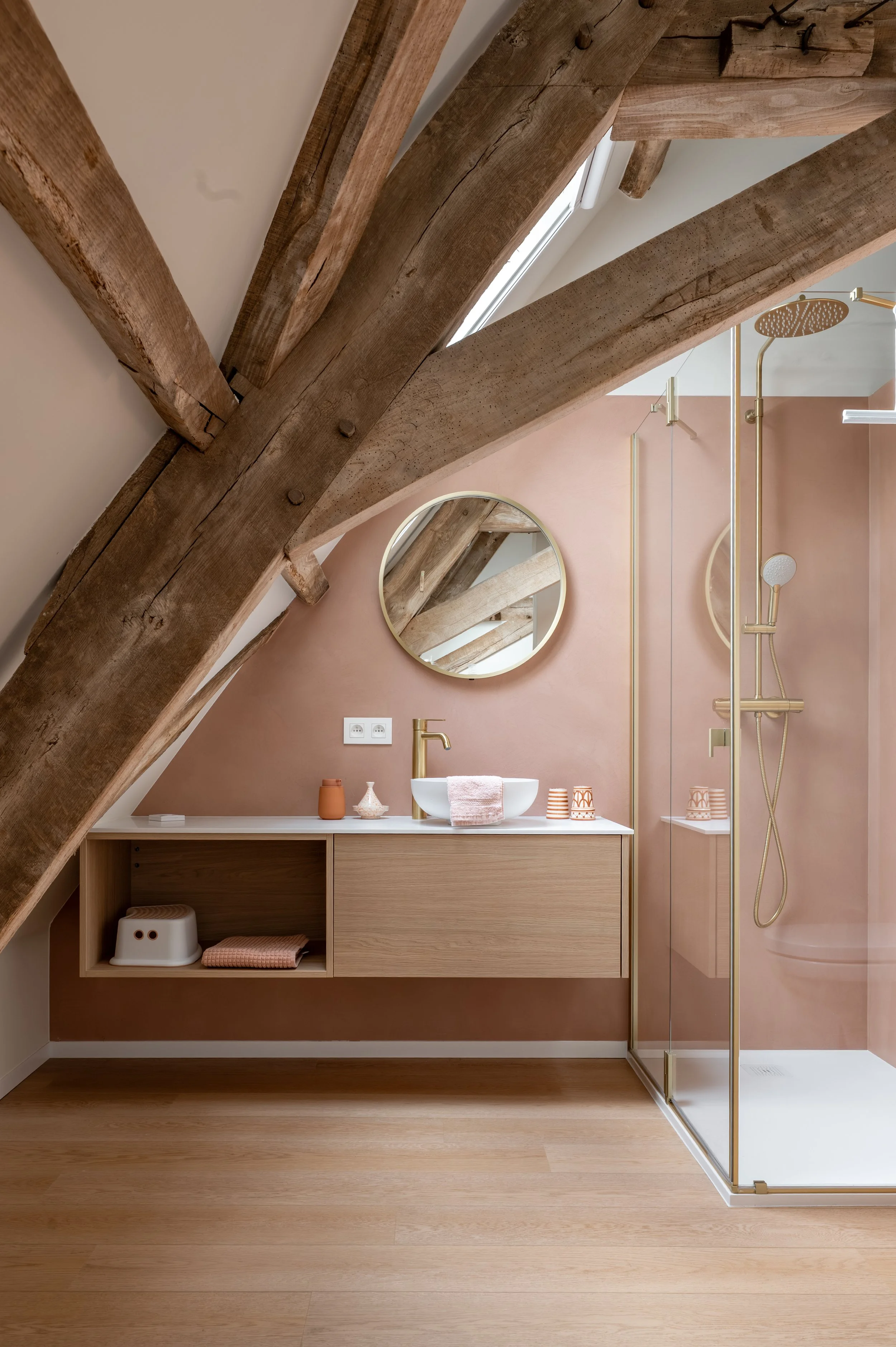 A modern bathroom with wooden beams on the ceiling, a peach-colored wall, and a floating wooden vanity with two shelves. The vanity has a white round vessel sink with a gold faucet. There are orange ceramic cups, a small vase, and a pink towel on the