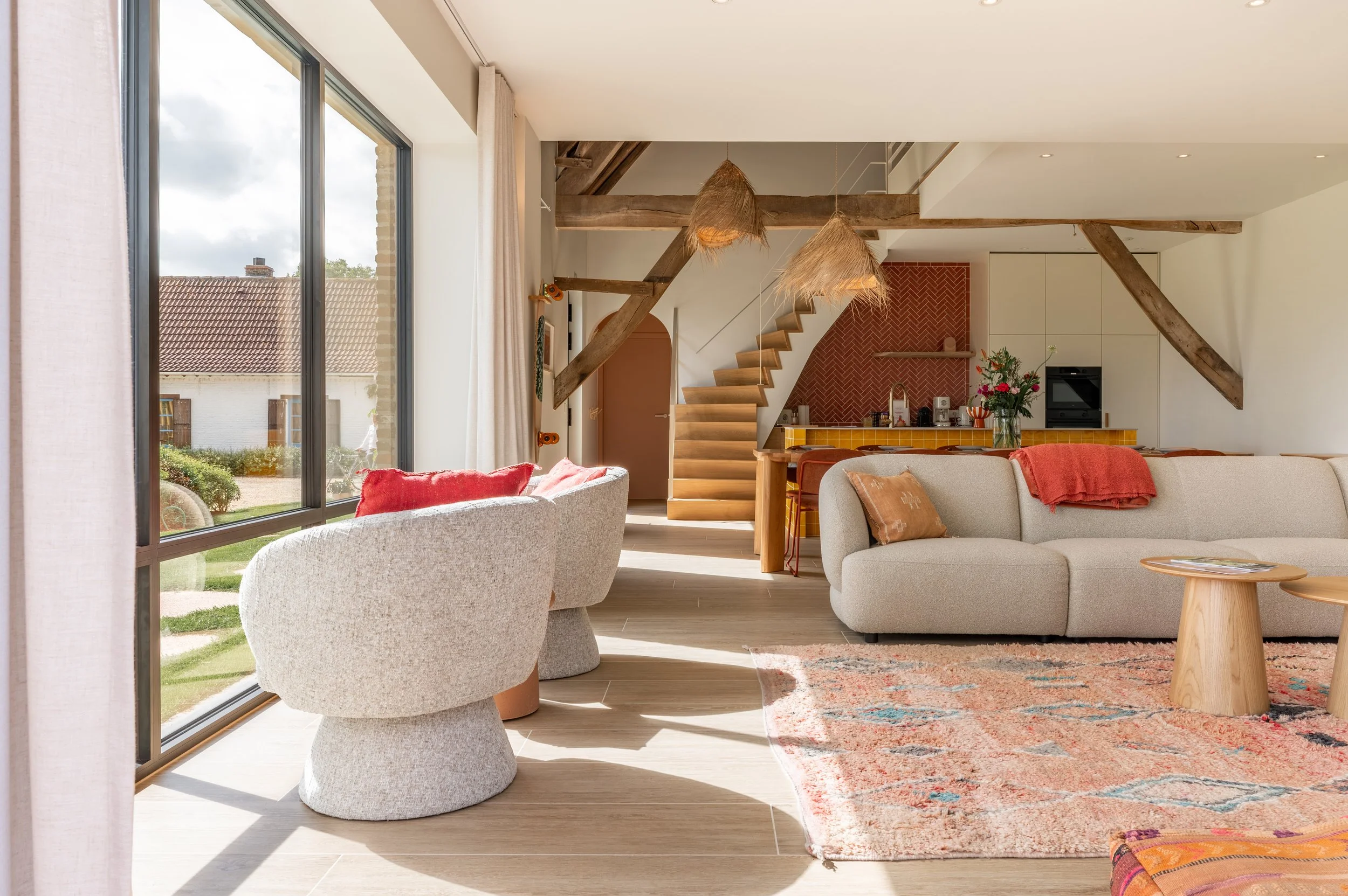 Bright living room with large windows, beige sofas, a colorful rug, and wooden beams on the ceiling.