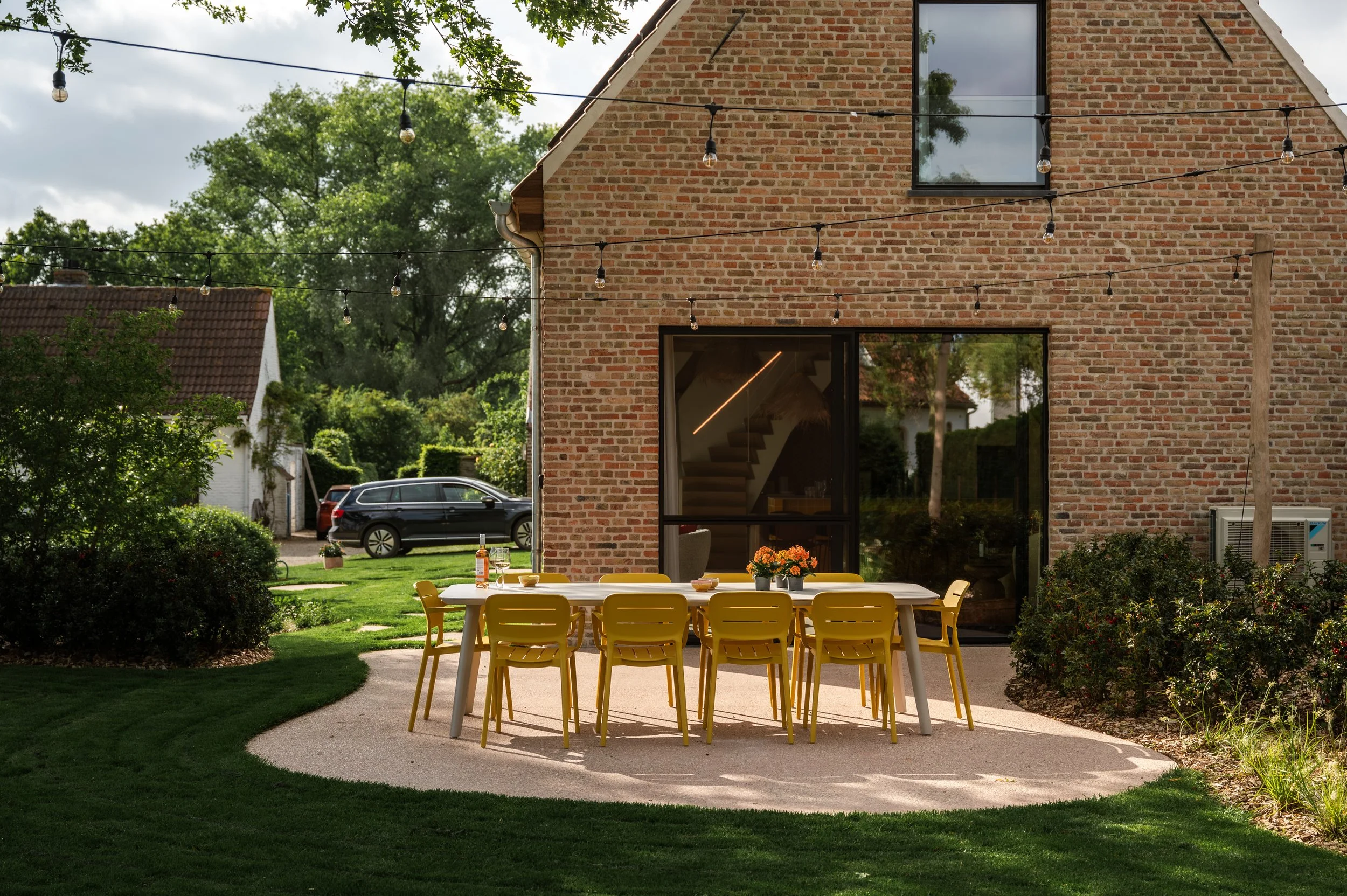 A backyard patio with a yellow table and matching chairs, overlooking a lush green garden with trees, bushes, and a brick house with string lights hanging overhead.
