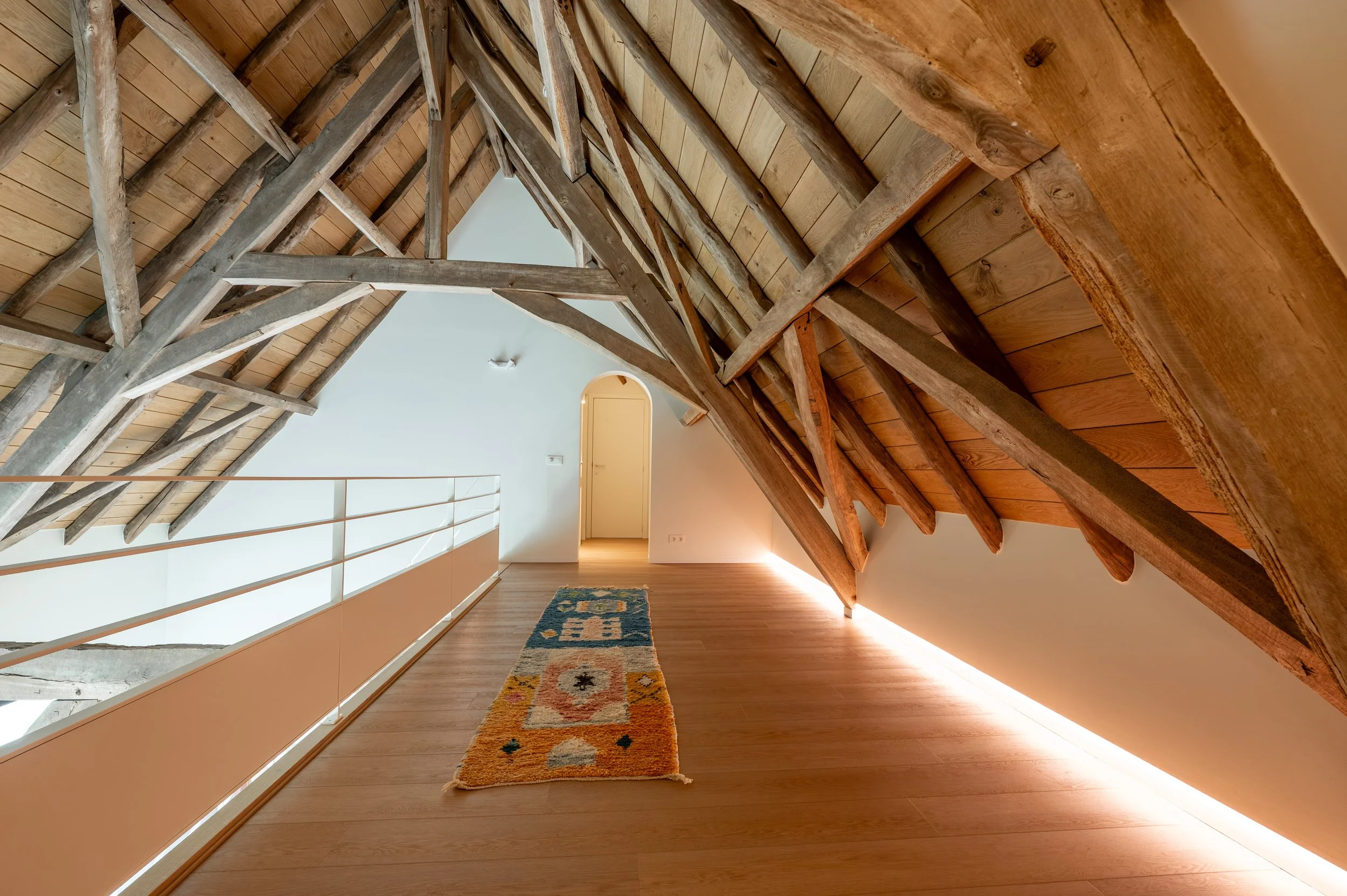 Interior view of an attic with wooden ceiling beams and a sloped ceiling, featuring a corridor with a colorful rug on a wooden floor and a railing along one side, leading to a door at the end.