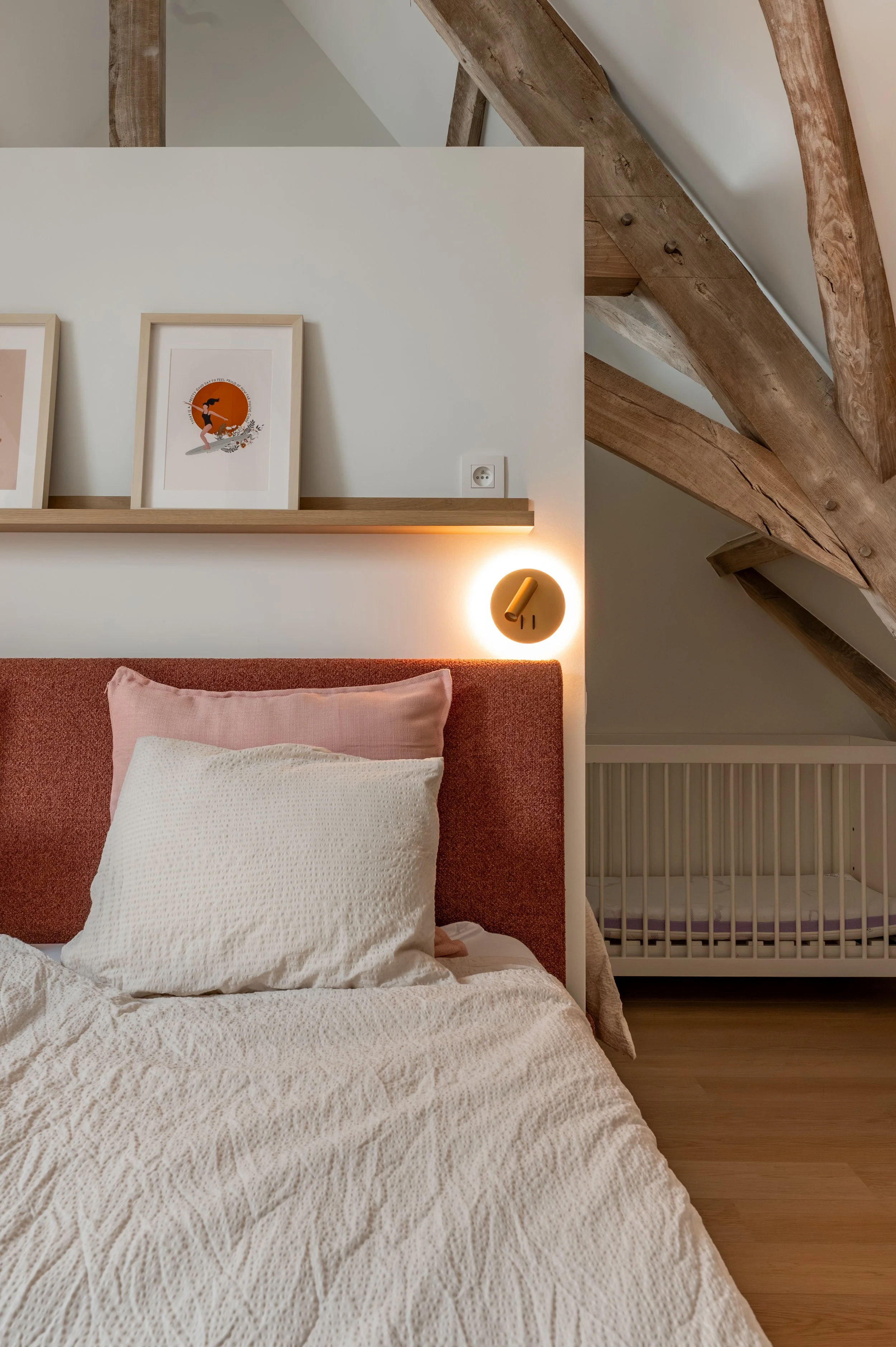 A bedroom with a bed that has white bedding and a pink pillow, a wooden headboard, framed artwork on the wall, a modern wall lamp, exposed wooden beams on the ceiling, and a baby crib in the corner.