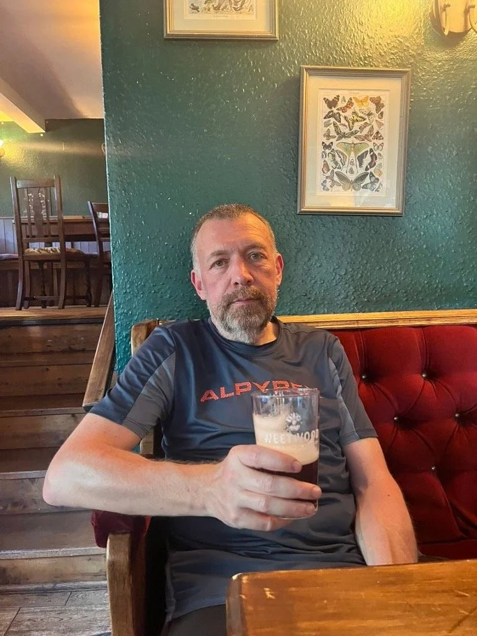 A man with a beard and short hair, wearing a dark athletic shirt, is sitting in a restaurant or pub, holding a glass of beer.