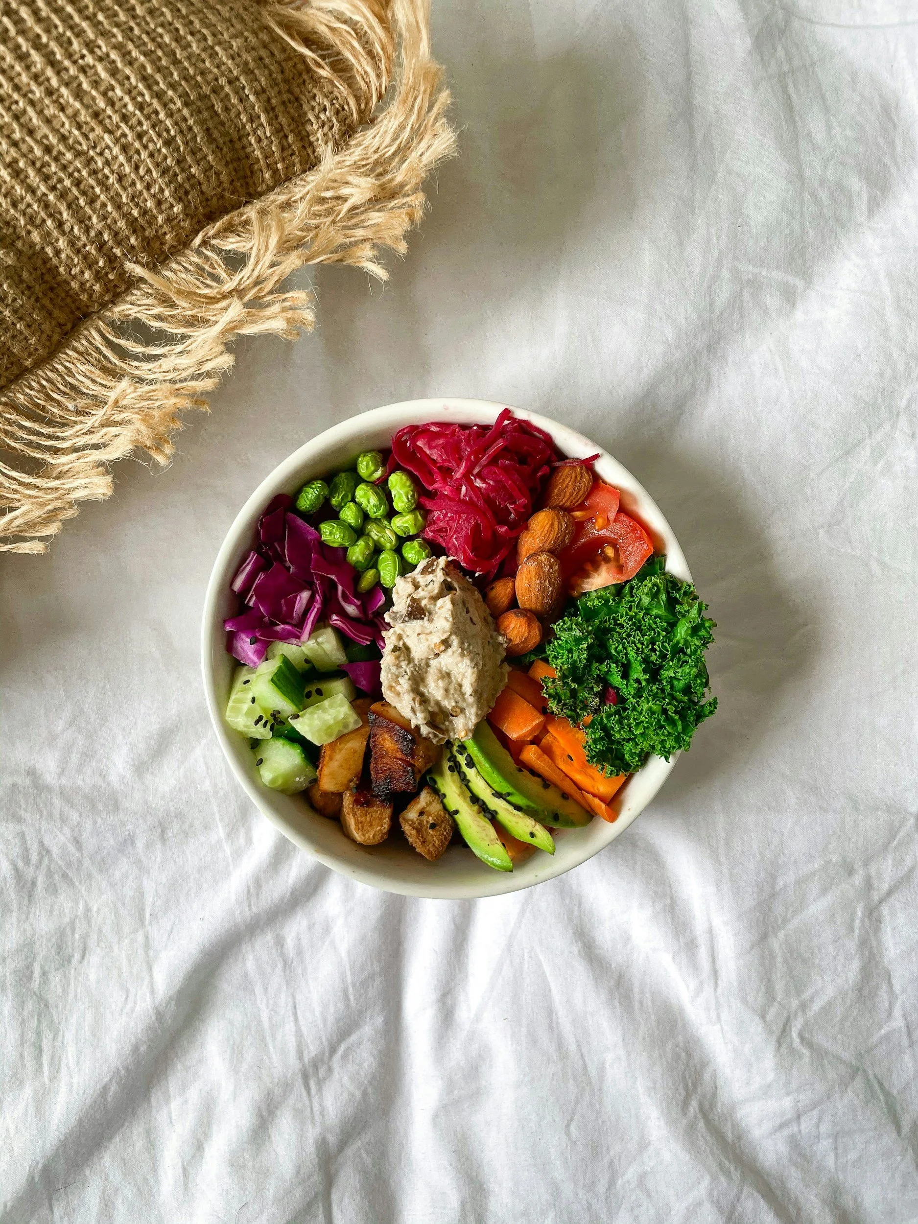 En bolle med ulike friske grønnsaker og nøtter, inkludert rødkål, agurk, cherrytomater, brokkoli, gulrøtter, avokado, kylling, hummus og bunashisablader, og et place av knuste nøtter på toppen.