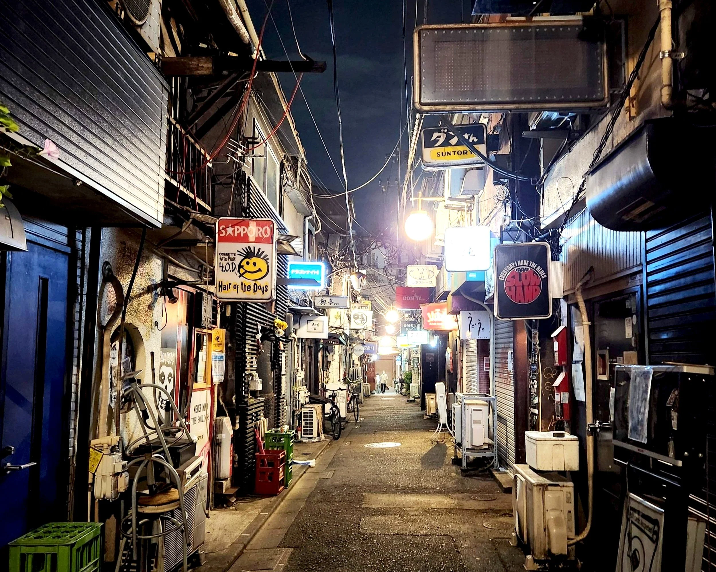 Night view of a narrow, lively alleyway with various illuminated signs for shops and restaurants, surrounded by storefronts with closed shutters and miscellaneous items along the street.