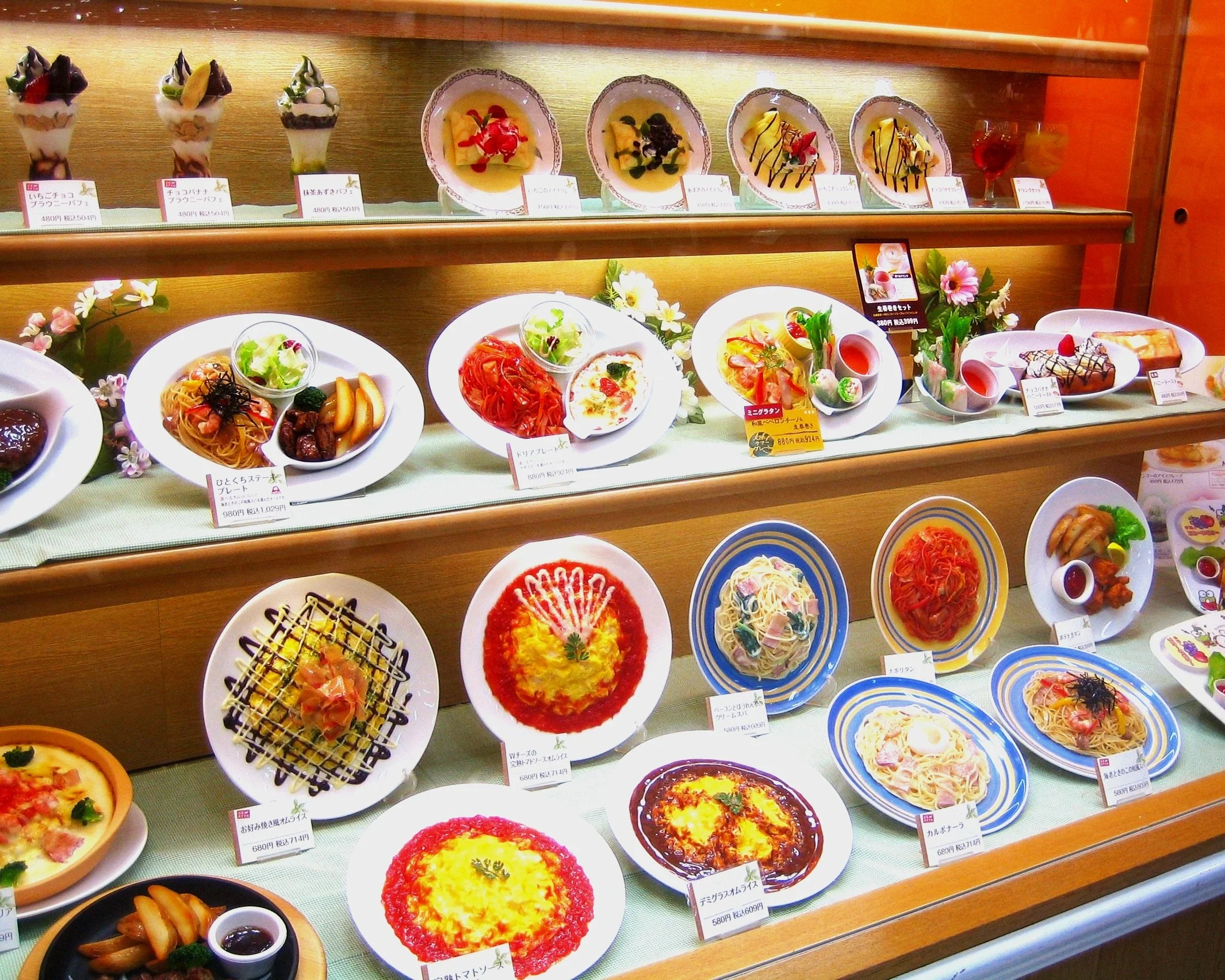 Display of various Japanese-style plastic food models, including desserts on the top shelf and plates of pasta, pizza, and salads on the lower shelves, arranged in a restaurant showcase.