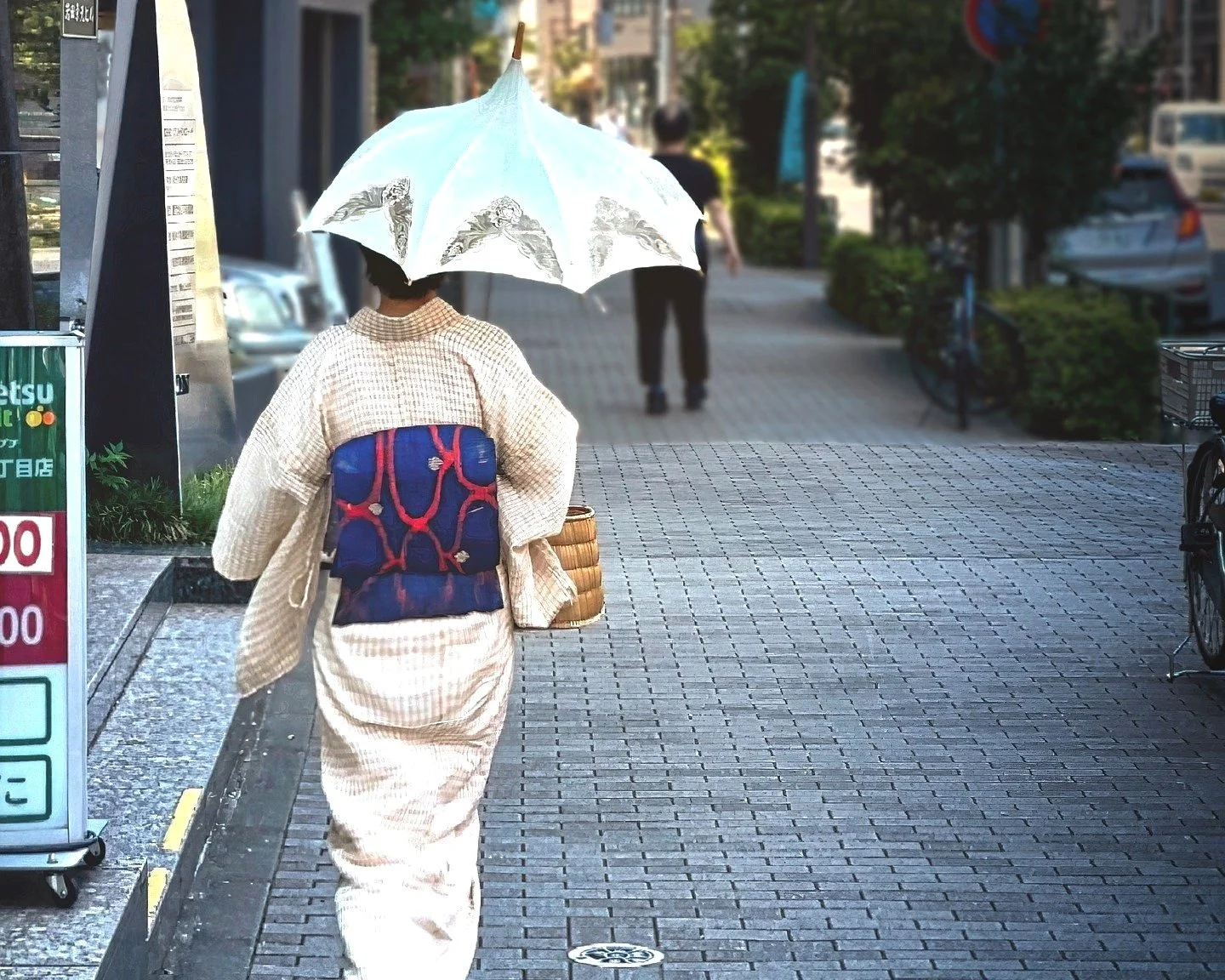 Woman wearing a kimono walking on a city sidewalk, holding a parasol with a traditional Japanese pattern, carrying a woven basket, with parked cars and bushes in the background.