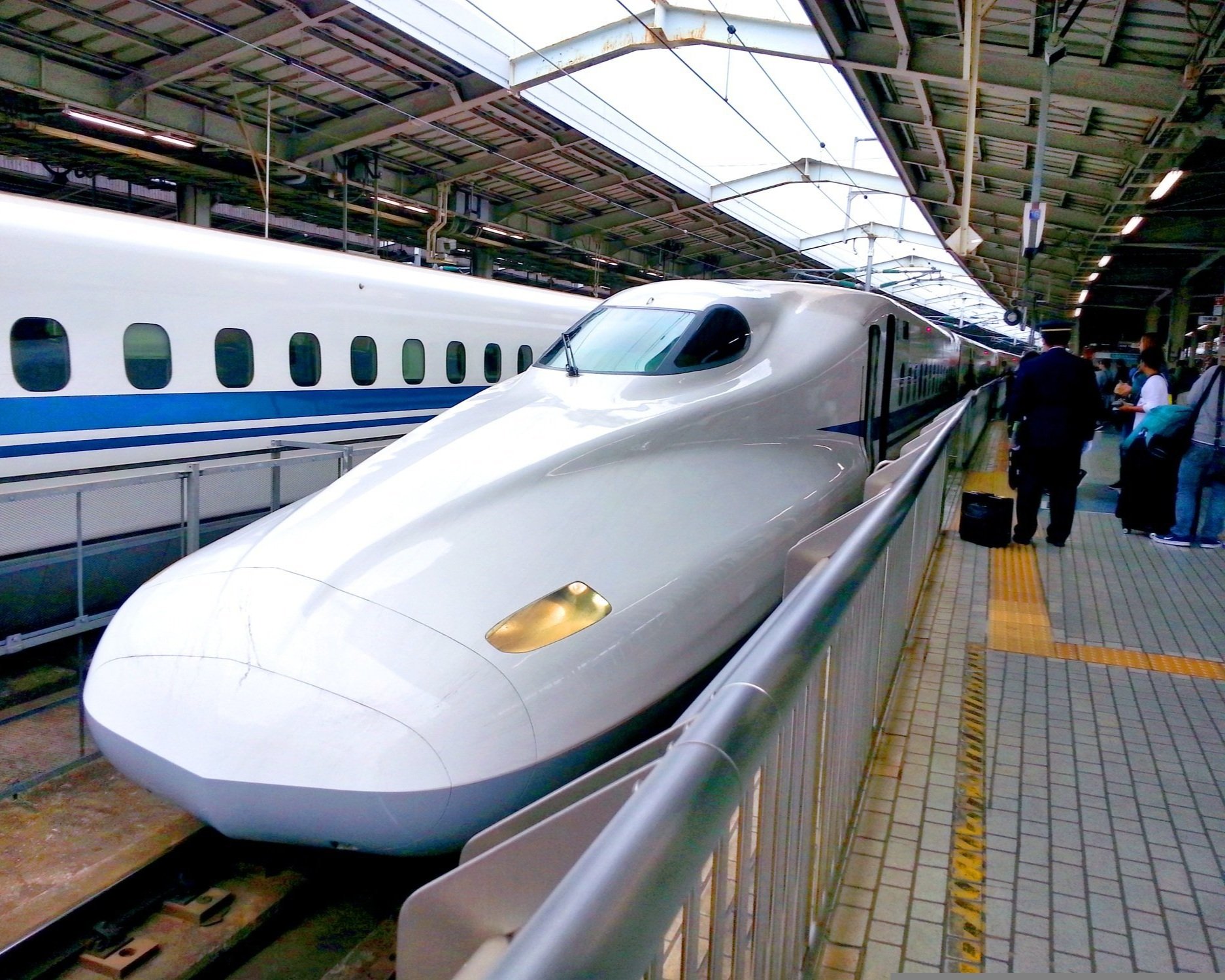 A sleek, white bullet train with a streamlined nose at a train station platform, with passengers nearby and another train visible in the background.