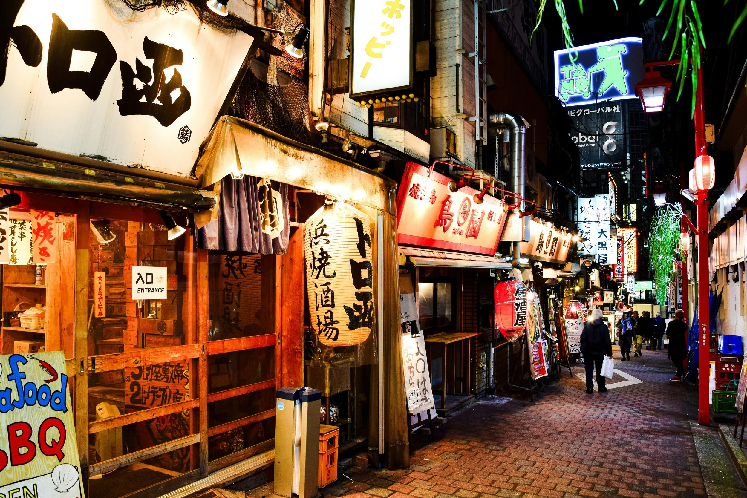 A bustling nighttime street scene with illuminated signs and storefronts in Japan, featuring traditional Japanese lanterns and colorful signs advertising food and drinks.