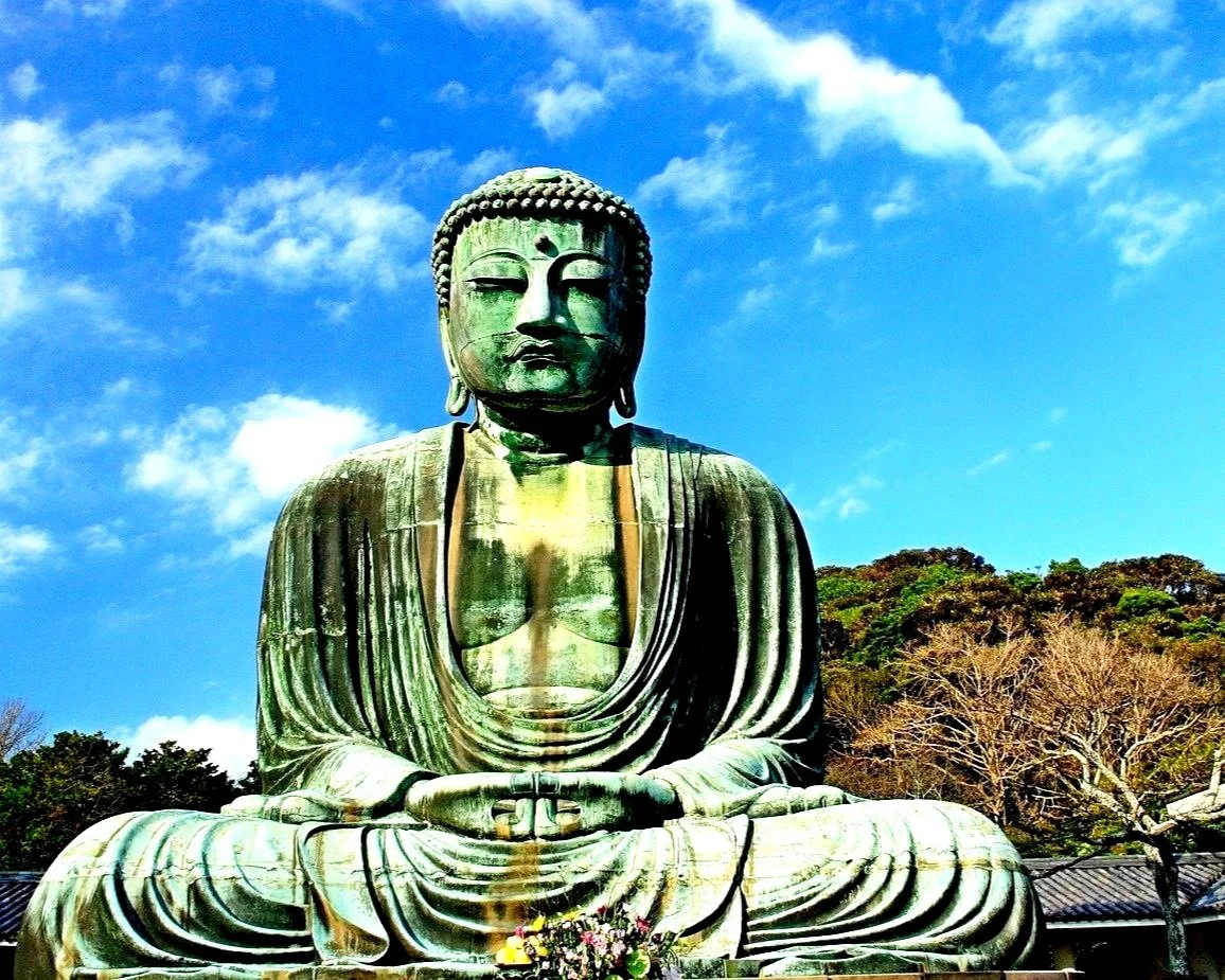 Large bronze statue of Buddha meditating outdoors under a blue sky with clouds, surrounded by trees.