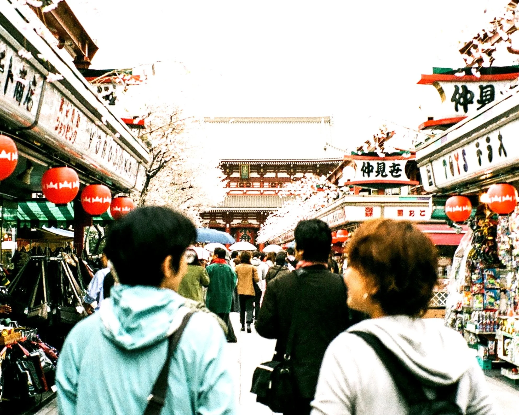 Crowd walking through a busy Japanese marketplace with traditional shops, red lanterns, and a temple gate in the background.