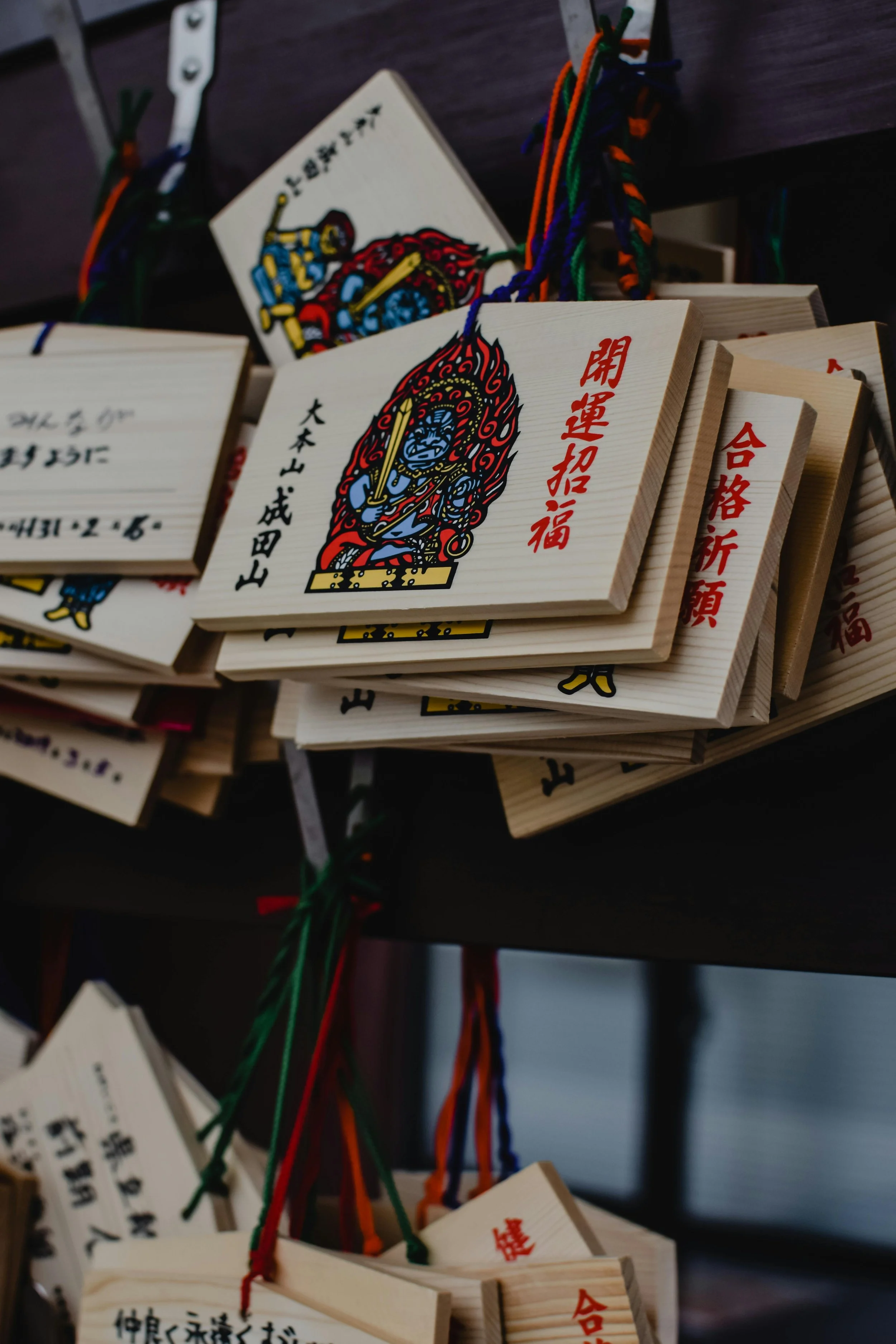 Wooden plaques with colorful illustrations and Japanese text hanging from multicolored strings.