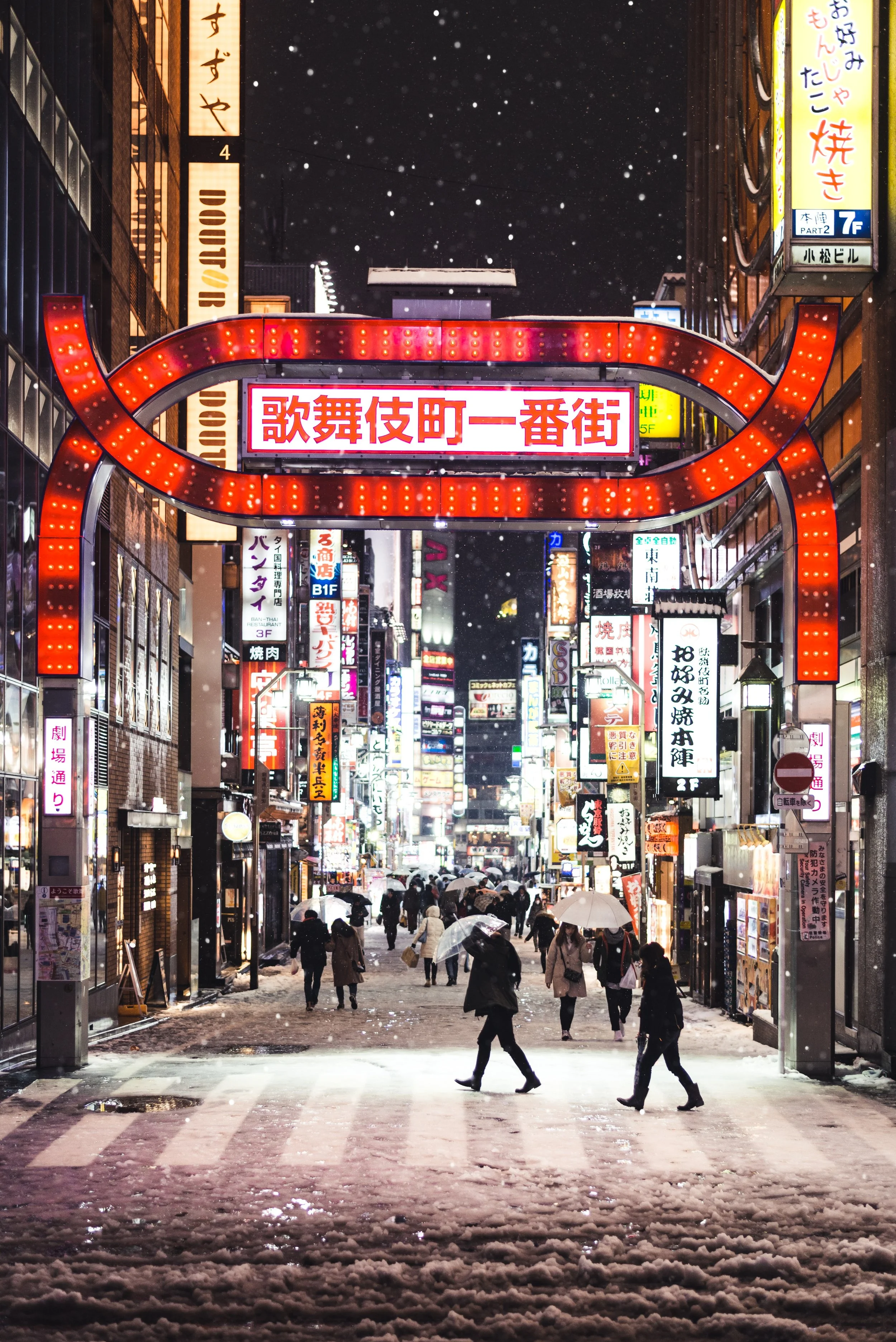 A busy city street in Japan at night with snow falling. The street is filled with illuminated signs in Japanese, and people holding umbrellas walk across the snow-covered crosswalk.