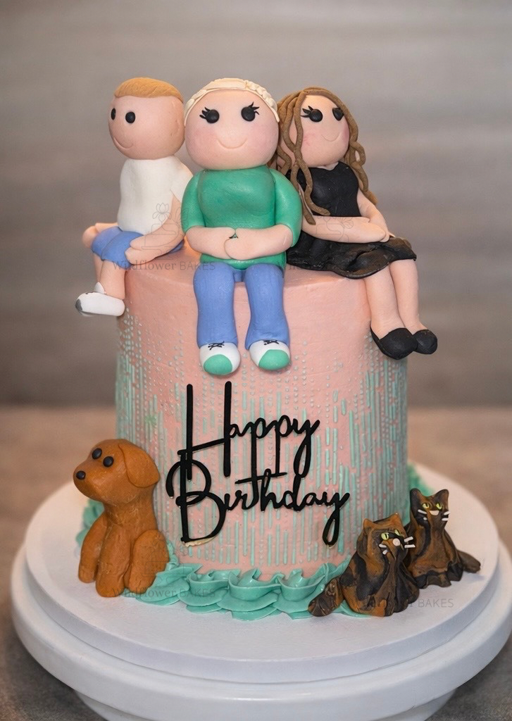 A pink birthday cake decorated with fondant figurines of three people sitting on top, a brown fondant dog, and two fondant cats, with a black "Happy Birthday" topper.