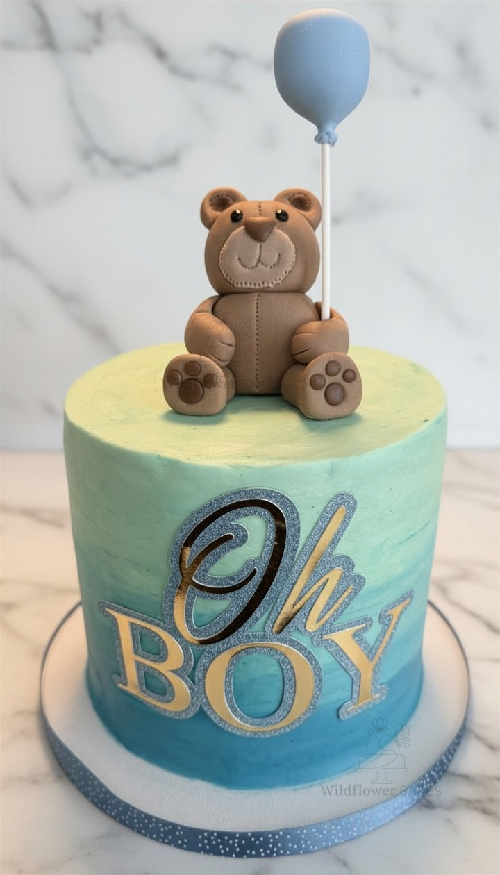 A cake decorated with a teddy bear holding a blue balloon and the words "Oh Boy" in silver letters.