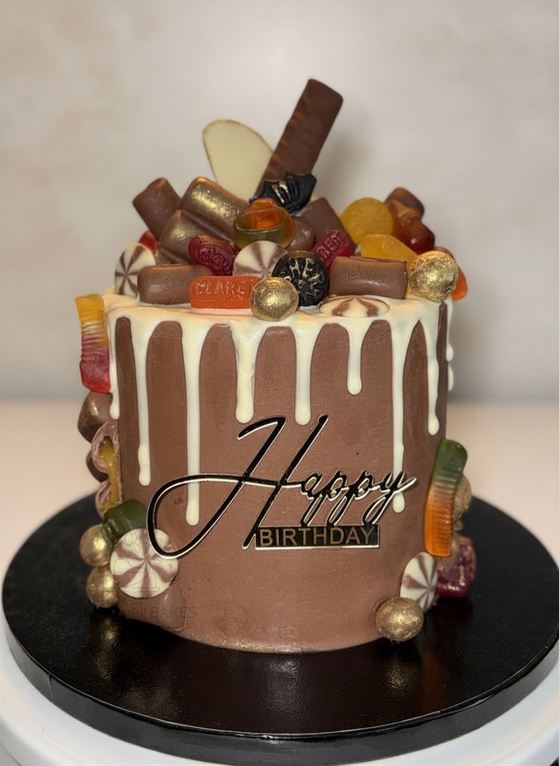 Chocolate birthday cake with white drip icing, topped with assorted candies, chocolates, and gold-colored decorations, and a "Happy Birthday" sign on the front.