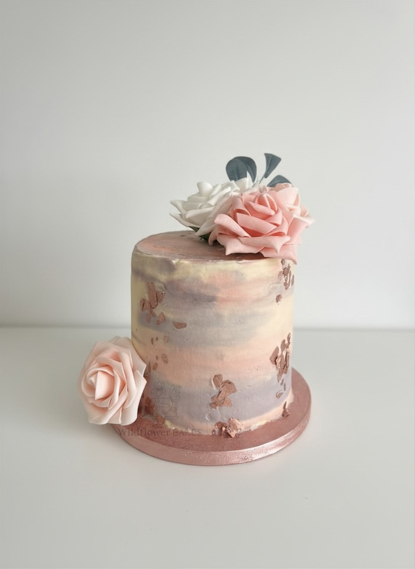 A tall, cylindrical marbled cake with pastel pink, white, and purple hues. Decorated with edible flowers in pink and white with some flowers placed on top and one on the cake board.