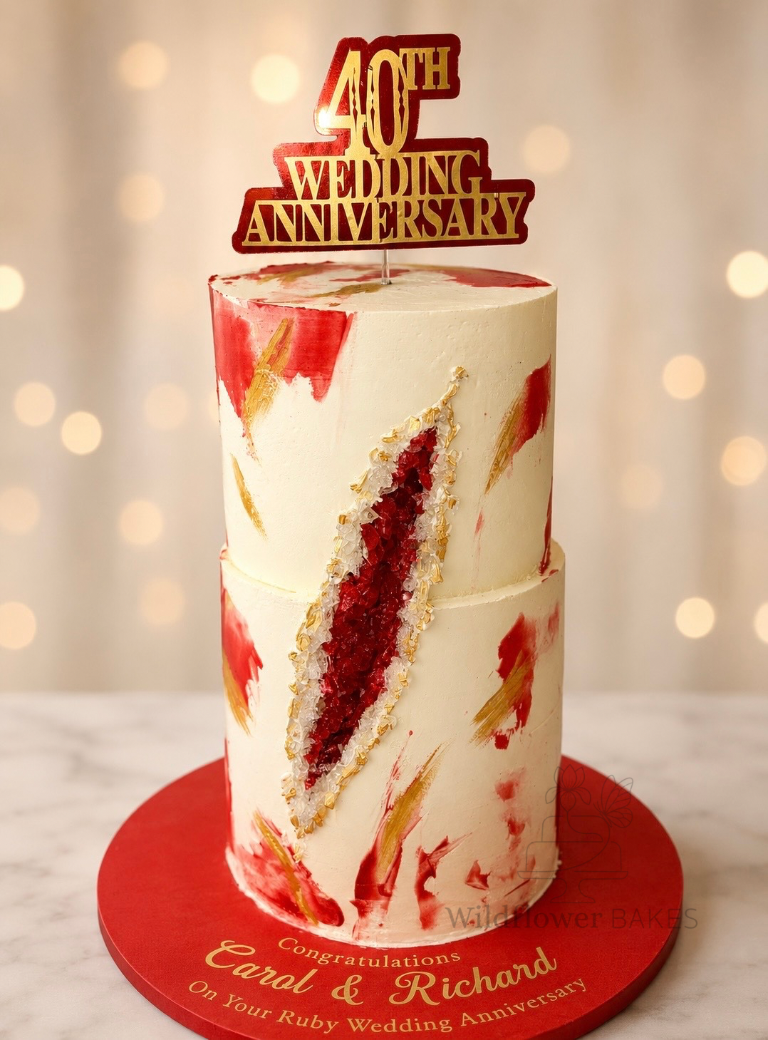 Two-tiered wedding anniversary cake with red, white, and gold decorative brushstrokes, topped with a '40th Wedding Anniversary' topper, placed on a red circular cake board with gold text celebrating Carol and Richard's ruby wedding anniversary.