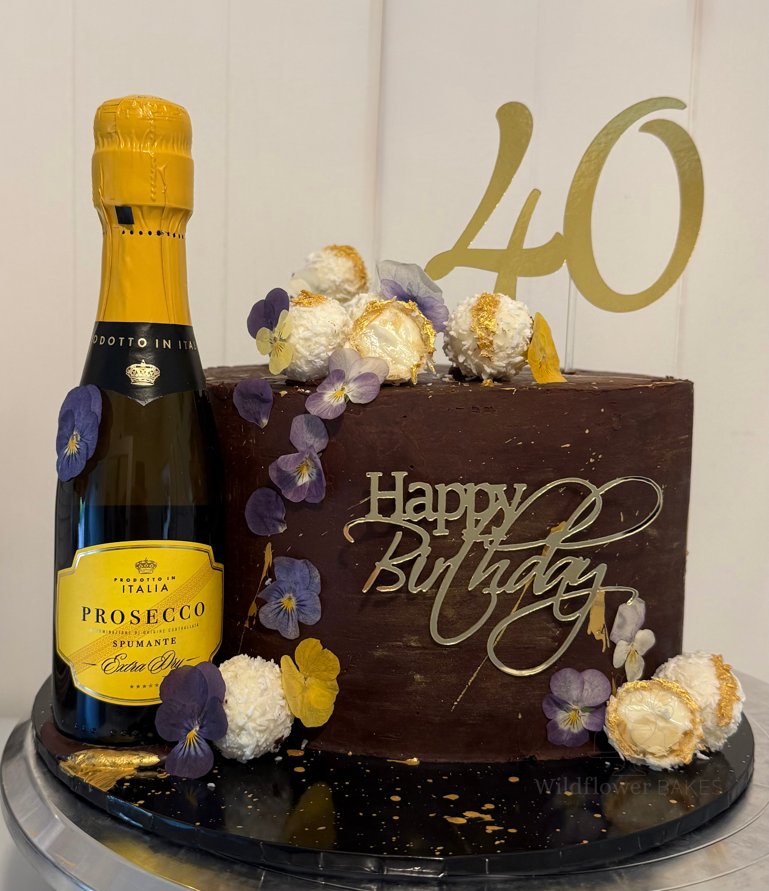A birthday cake with purple and yellow flower decorations, a bottle of Prosecco, and a gold "40" cake topper. The cake has white whipped cream and gold accents. The background features a white wall and a wooden panel with a gold "Happy Birthday" sign
