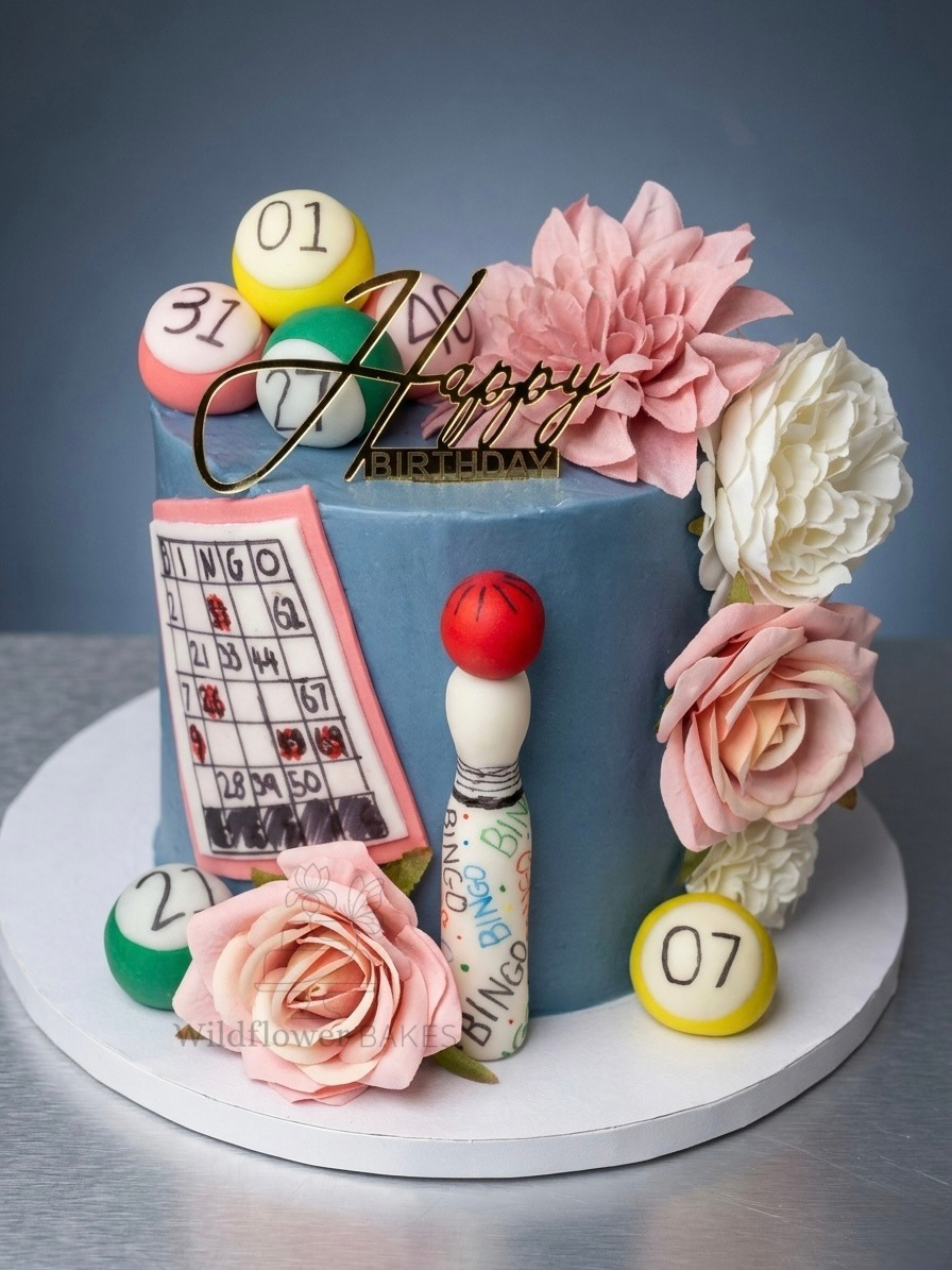 A blue birthday cake decorated with cotton candy pink and white flowers, bingo balls, a bingo card, a bingo dabber, and a gold 'Happy Birthday' cake topper.