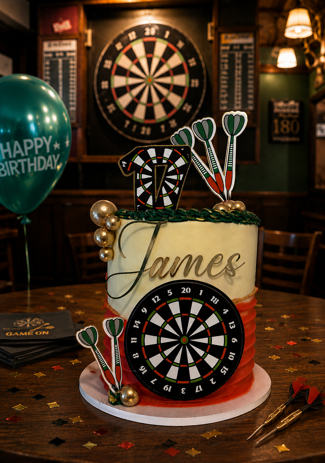 A birthday cake with dartboard decorations and the name 'James' on it, set on a wooden table with a green balloon and darts nearby, in a pub setting.