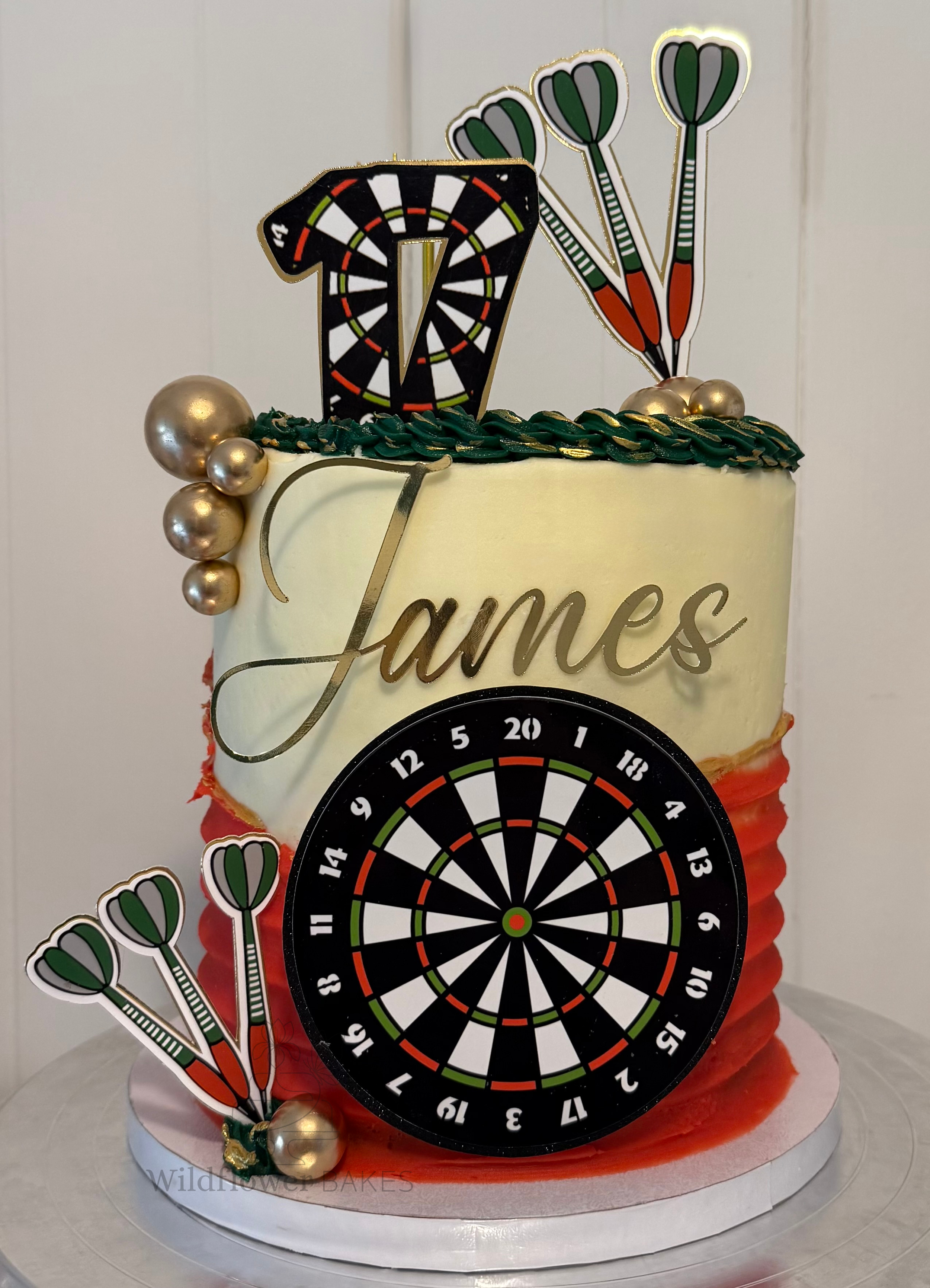 A birthday cake decorated with gold spheres, green and gold icing accents, and dartboard-themed toppers and decorations.