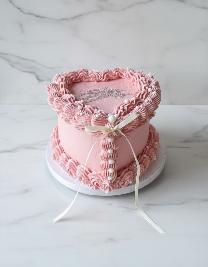 Pink heart-shaped cake decorated with pink buttercream frosting, white pearl-like icing, and a white ribbon bow.