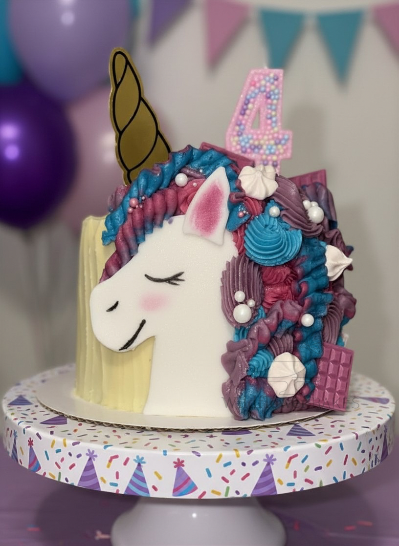 A unicorn-themed birthday cake with a white unicorn face, a colorful mane with blue, purple, and pink frosting, a golden horn, and pink ears. The cake is decorated with edible pearls and has a pink birthday candle shaped as the number 4. The cake is 