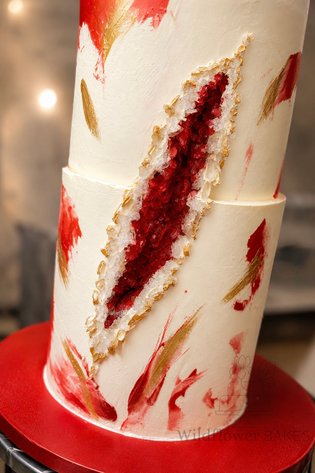 A tall, round, multi-layered cake with smooth white icing and red and gold abstract brush strokes. A section of the cake is cut out to reveal a red and white geode with golden accents. The cake is placed on a red cake board.