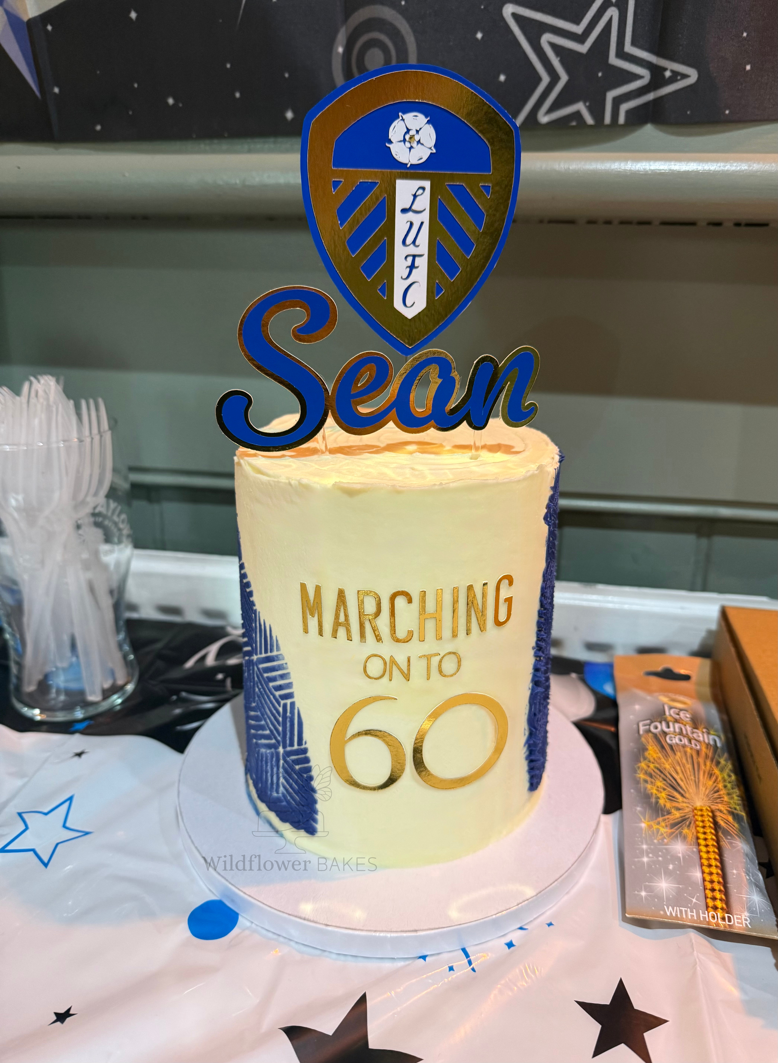 Cake decorated with a Leeds United badge cake topper, with the text 'Marching On To 60', positioned on a table with star-themed tablecloth, surrounded by party favors and decorations.