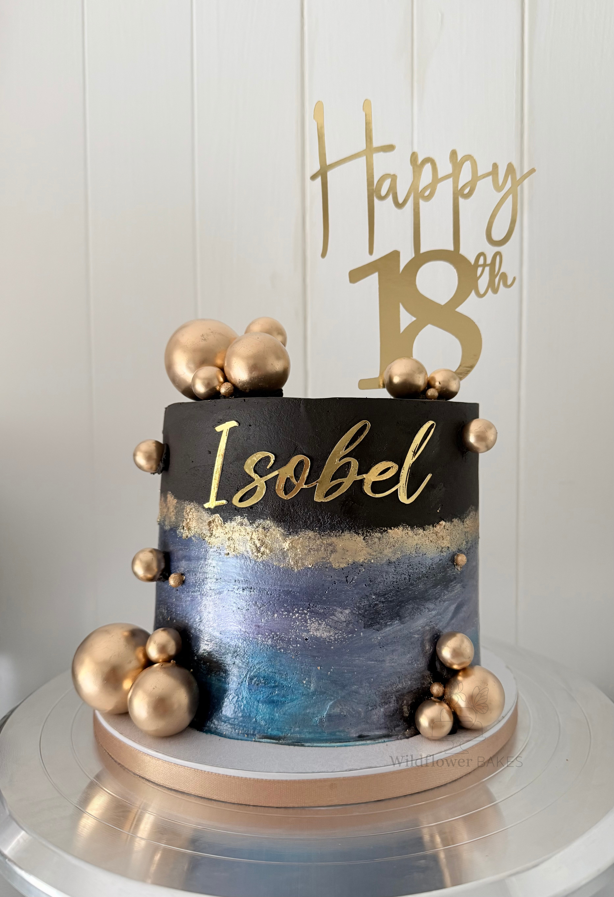 A black and silver birthday cake with gold lettering that reads "Isobel" and gold spherical decorations. A gold sign on top of the cake reads "Happy 18th."