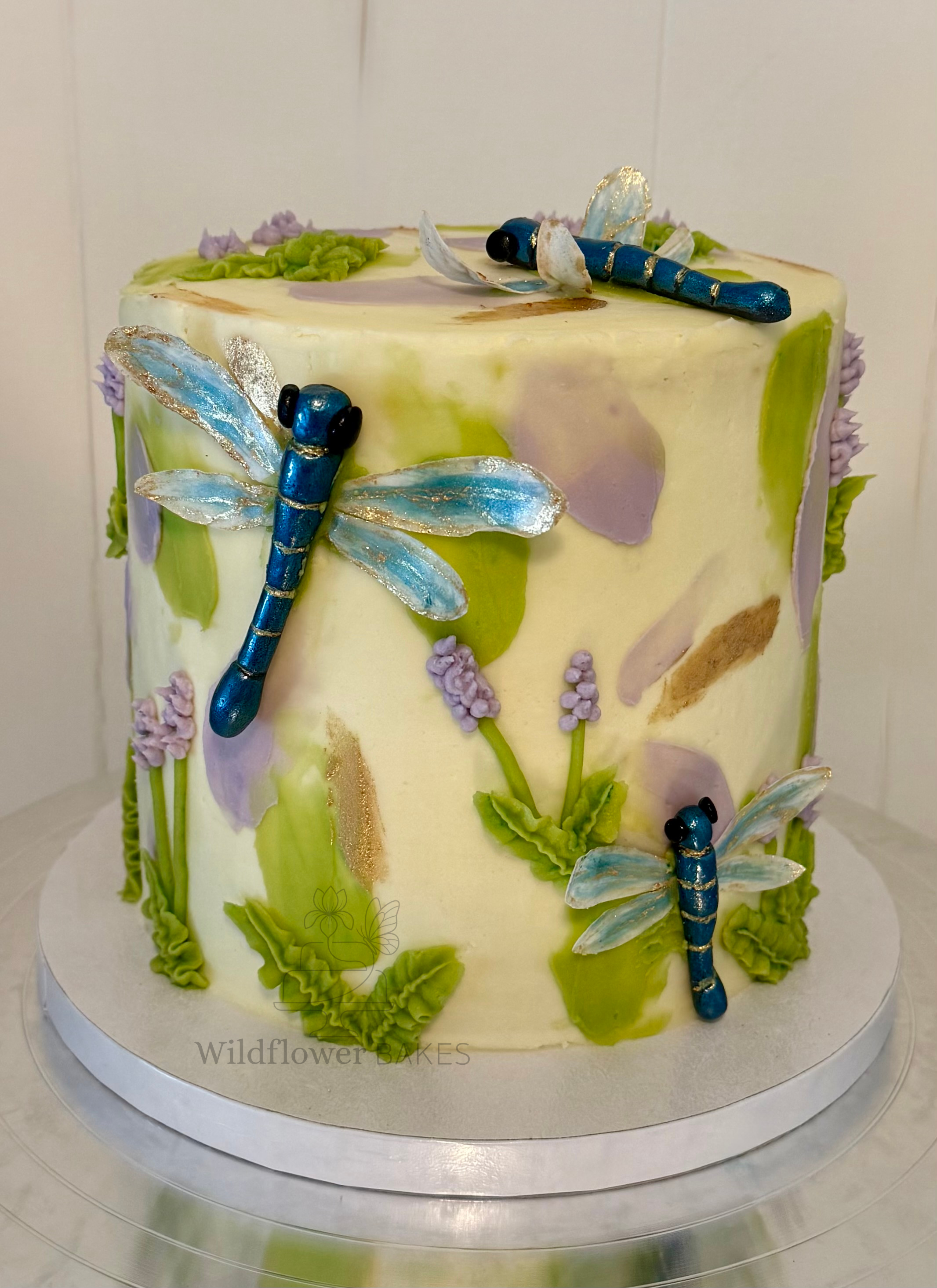 Decorative cake with edible blue and gold dragonflies, purple and green icing flowers, and green icing leaves and ruffles.