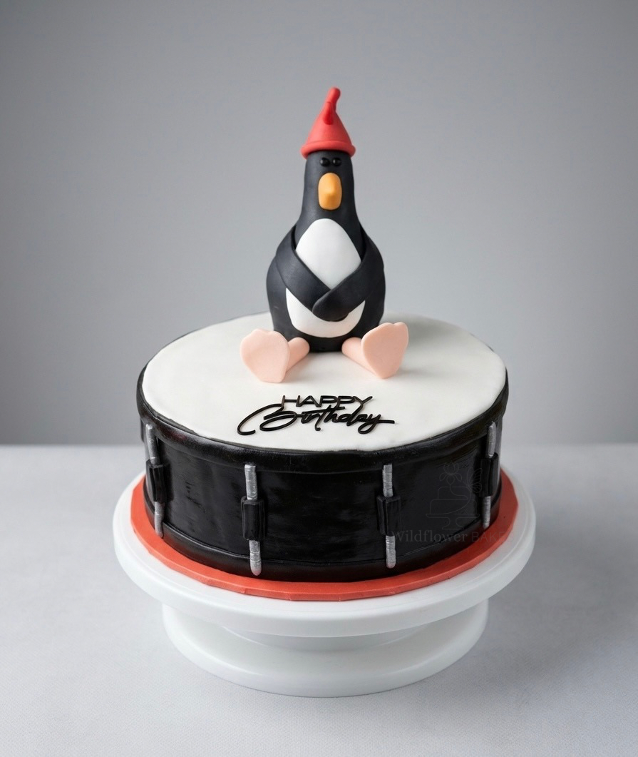 A birthday cake with an edible topper of Feathers McGraw, sitting on a white and black cake decorated with a drum design and the words "Happy Birthday" written on top.