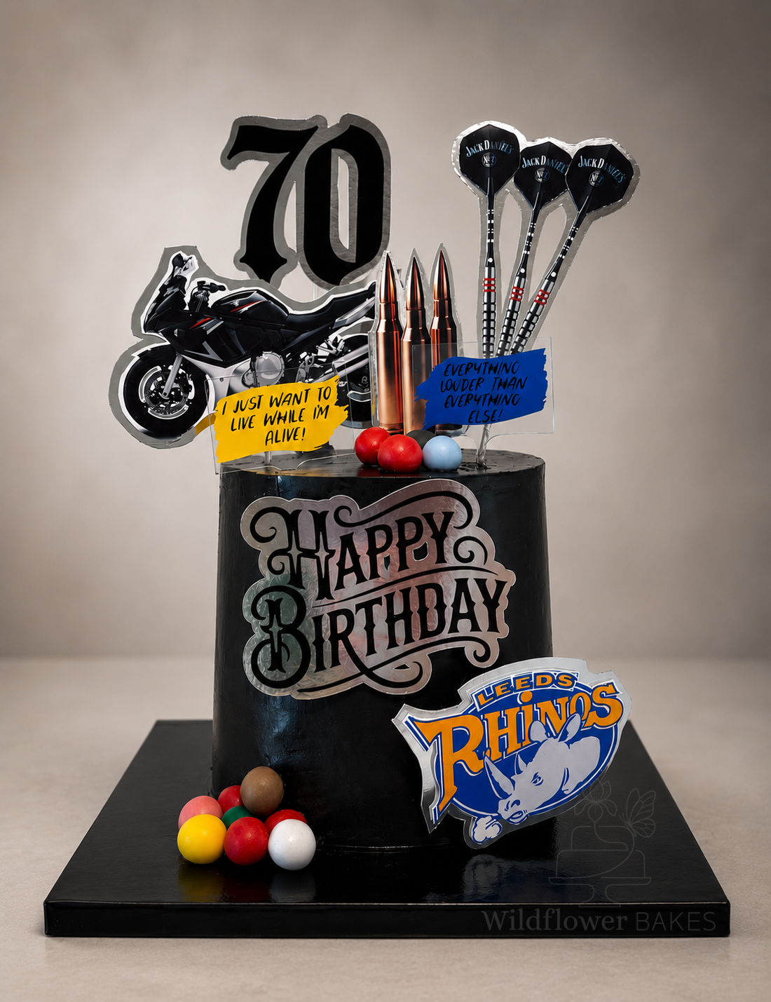 Black birthday cake decorated with aviation and sports items, including a motorcycle cutout, darts, and colorful balls. The cake features a large 'Happy Birthday' message, a '70' sign, and signs with humorous quotes. It also has a 'Leeds Rhinos' logo