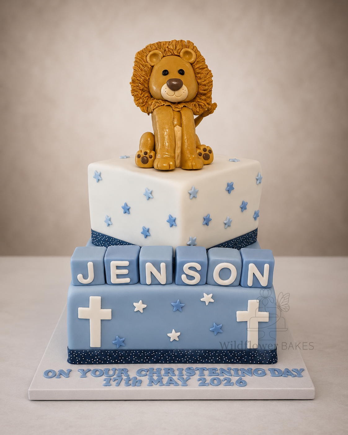 A Christening Day cake with a lion figurine on top, decorated with blue stars and a ribbon, featuring the name Jenson and the message "On your Christening Day, 17th May 2026".