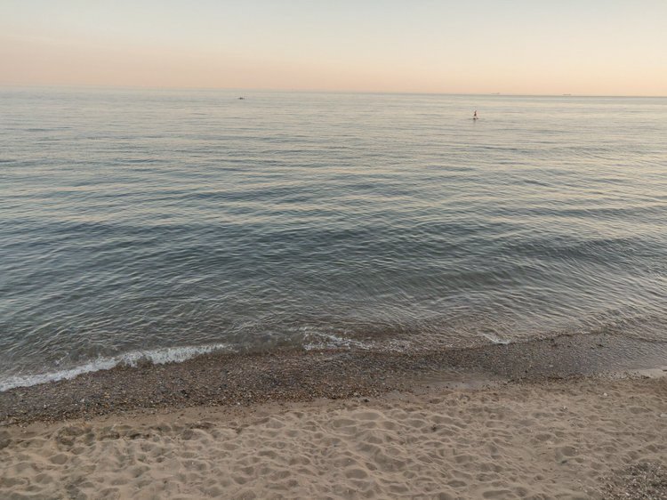 Calm ocean water near a sandy beach at sunset with a lone paddleboarder in the distance.