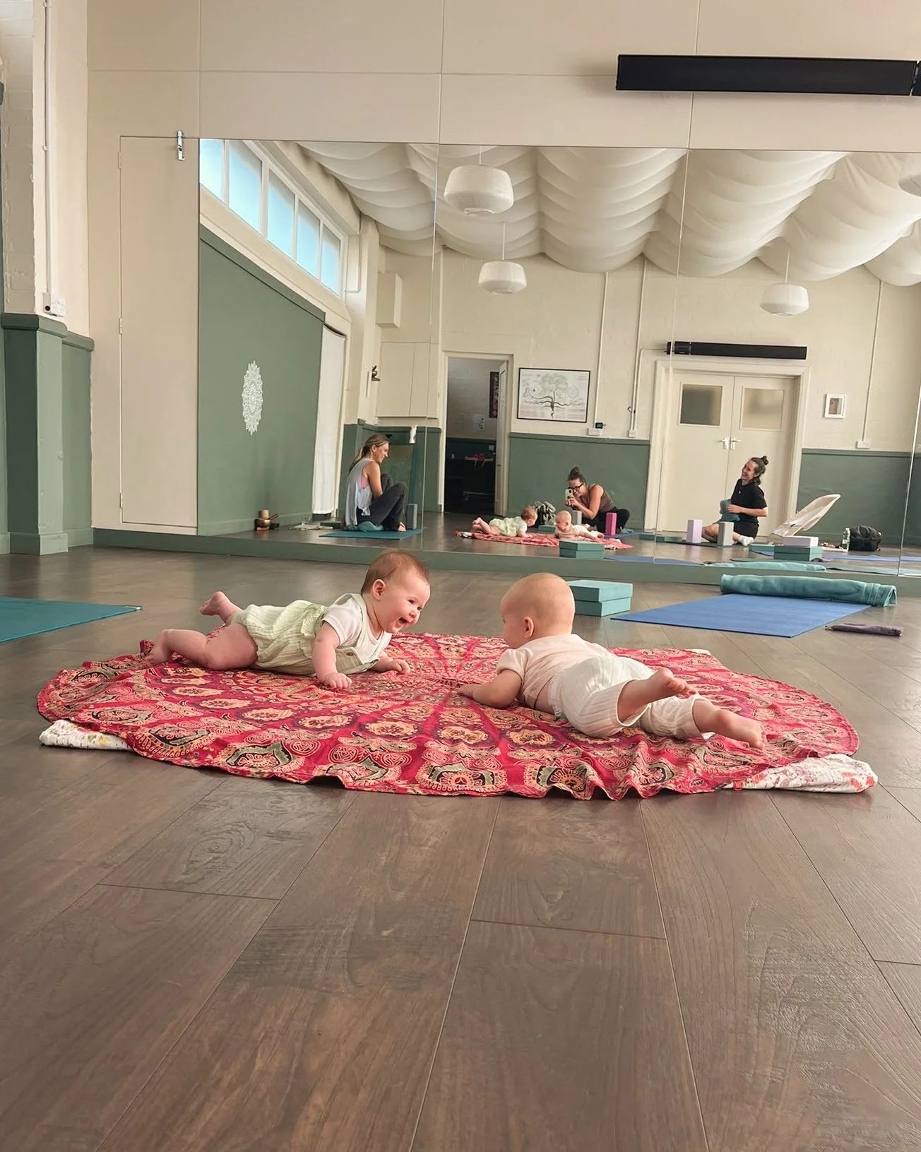 Babies on a mandala mat, in a studio with a yoga mat with props behind them and yoga students and teacher reflected in the mirror