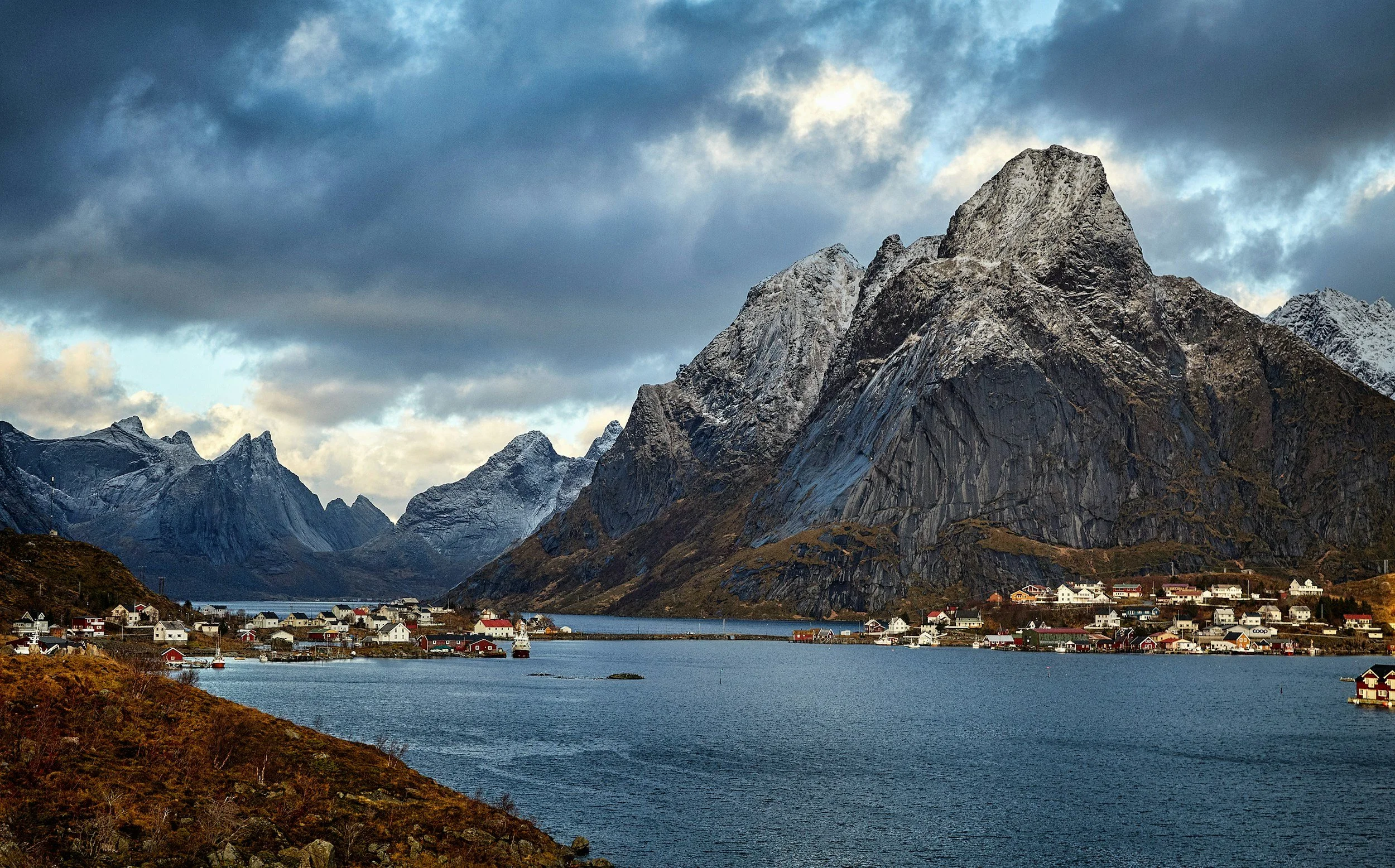 Norway, a landscape view of a coastal village with colorful houses at the foot of tall, snow-capped mountains under a cloudy sky, with water in the foreground.