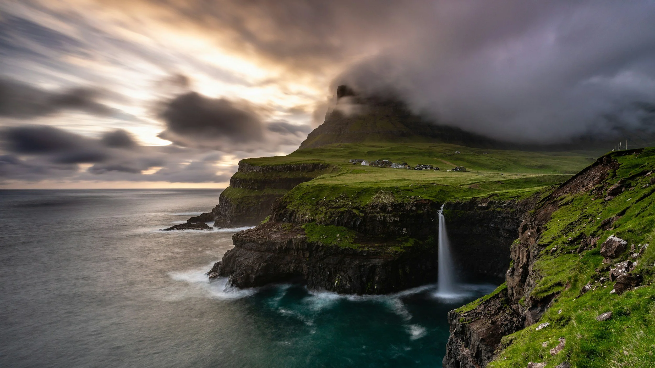 The Faroe Icelands, with cliffs overlooking the ocean and a waterfall cascading down the rocks, green grass covering the cliff top, small houses and a mountain shrouded in clouds or fog in the background.