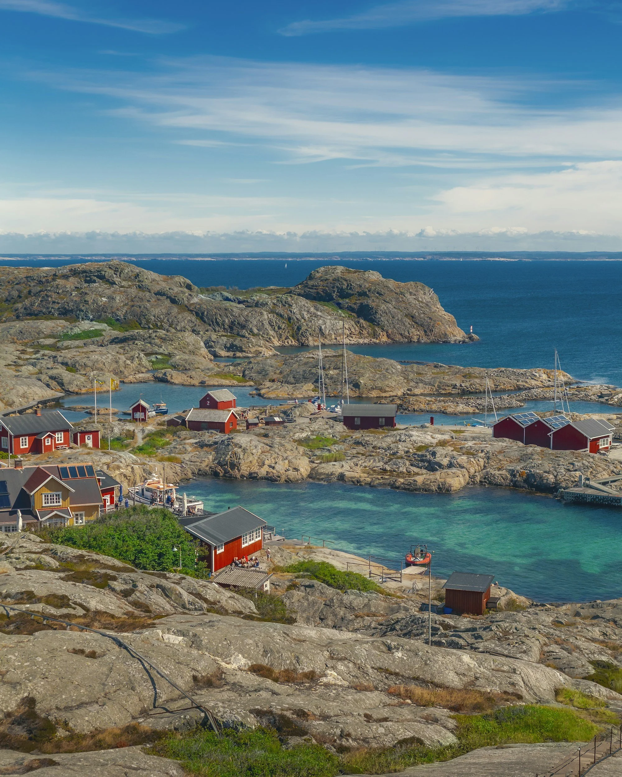 Sweden, a coastal scenery with rocky shoreline, small boats, red houses, and calm blue water under a partly cloudy sky.