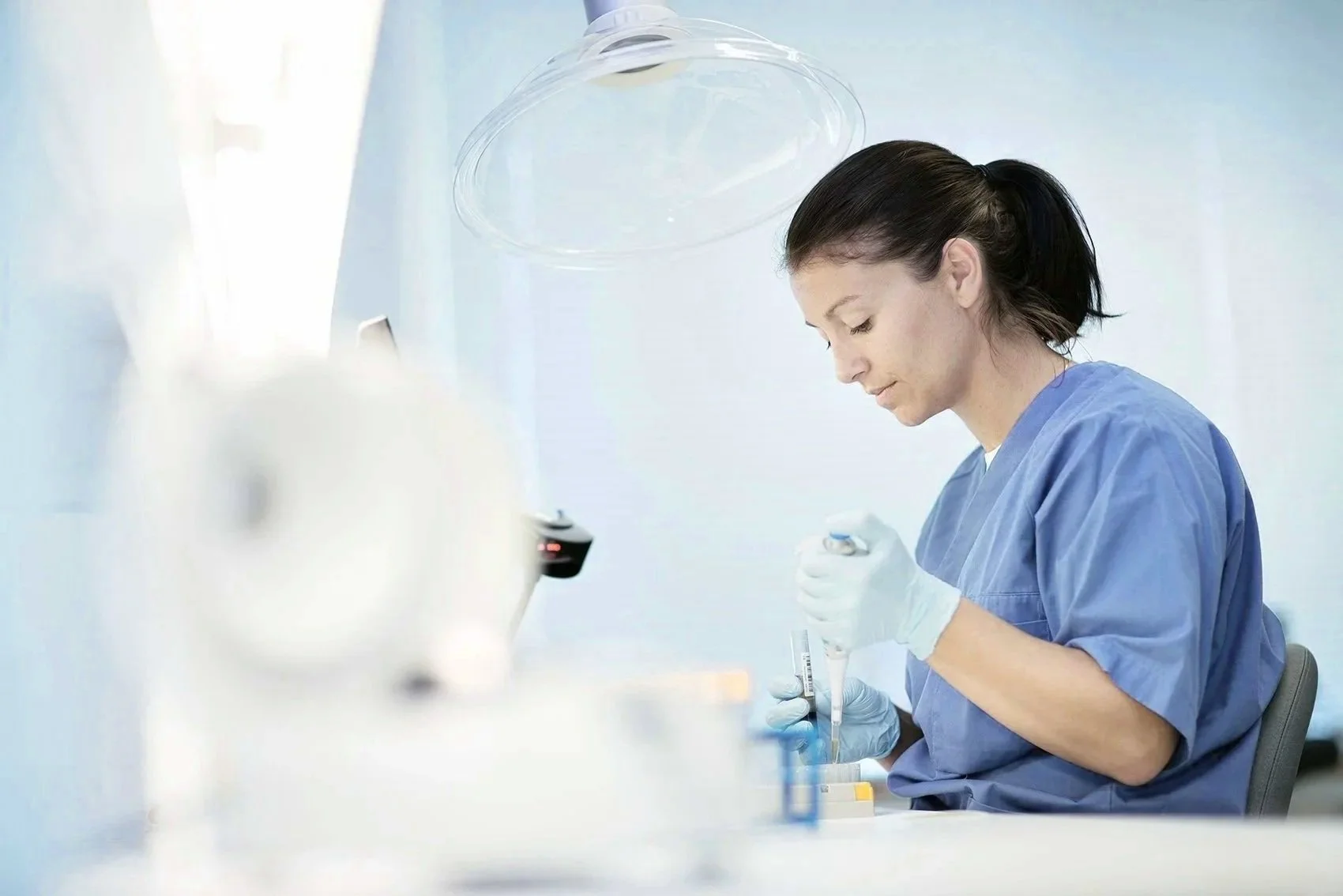 A female biomedical scientist wearing scrubs and gloves working with a pipette in a laboratory.