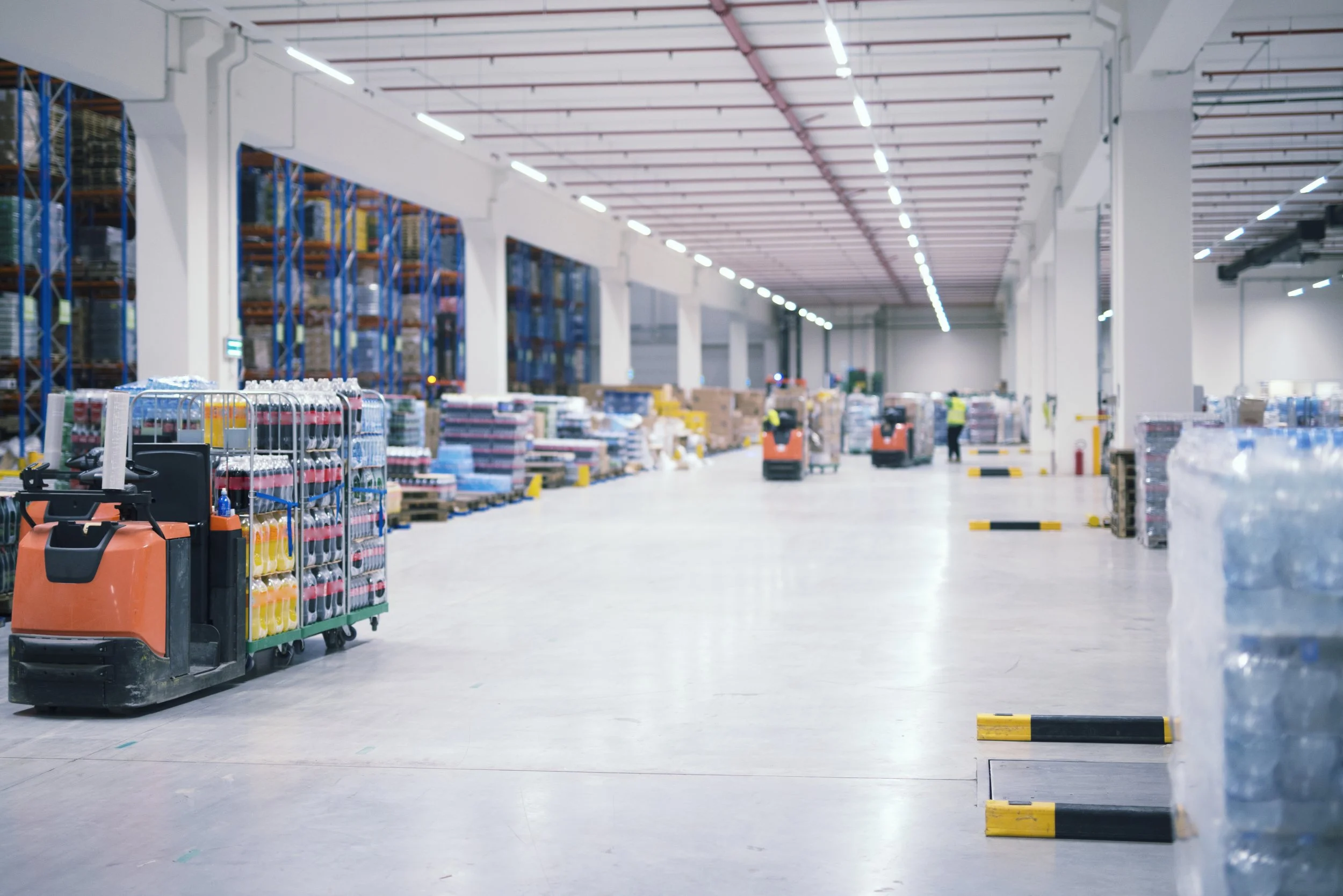 Image of a large, empty warehouse or distribution center with cluttered shelves on the left side. In the foreground, there are orange and black electric carts with supplies. In the background, there are workers, shelves, and equipment, with bright overhead lighting and an industrial ceiling.