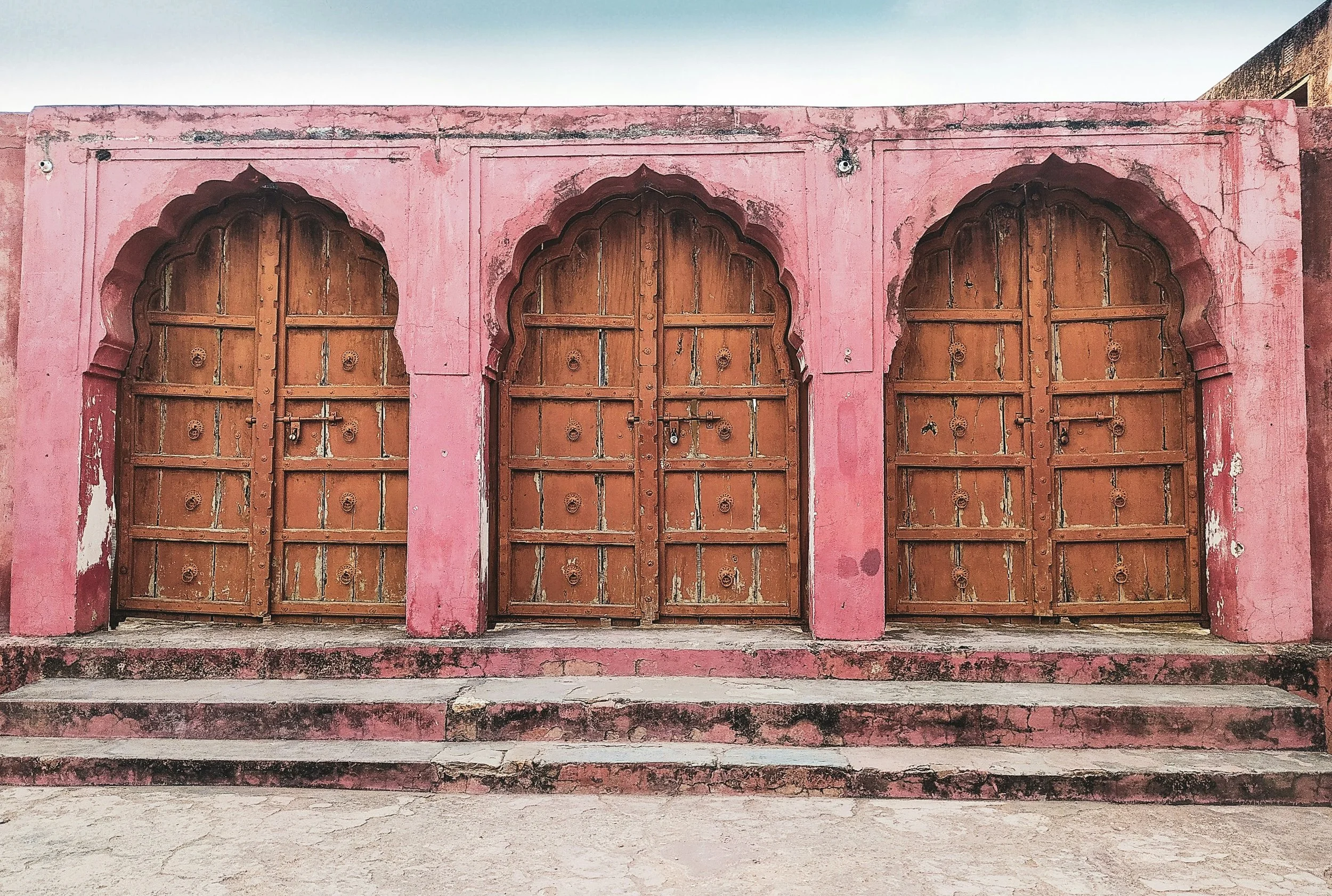 A pink wall with three large wooden, arched doors that are weathered and slightly open, with a small overhang above and steps leading up to the entrance.
