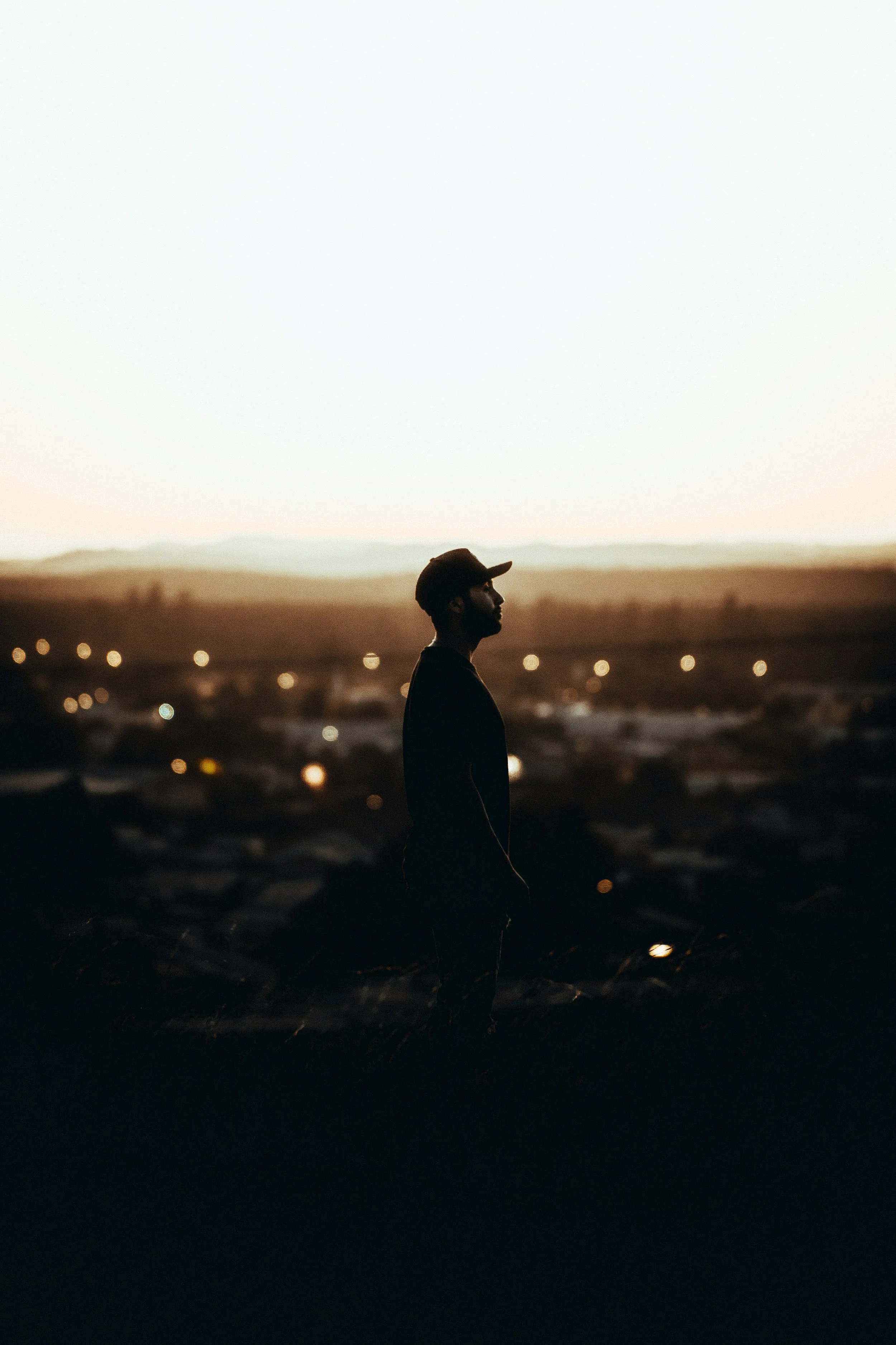 Silhouette of a man wearing a baseball cap standing outdoors during sunset or dusk, with a blurred background of lights and terrain.