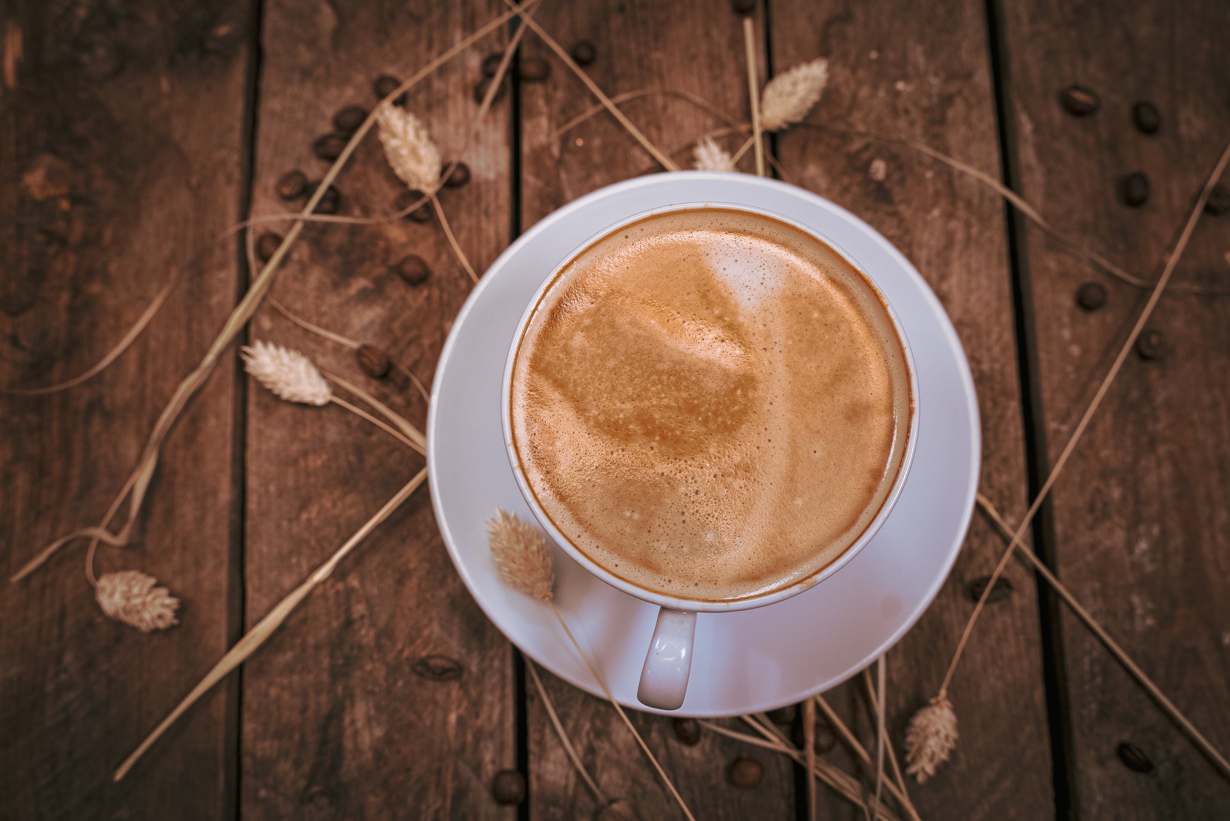 A white cup of coffee with frothy surface on a saucer, placed on a wooden table decorated with dried plants and coffee beans scattered around.